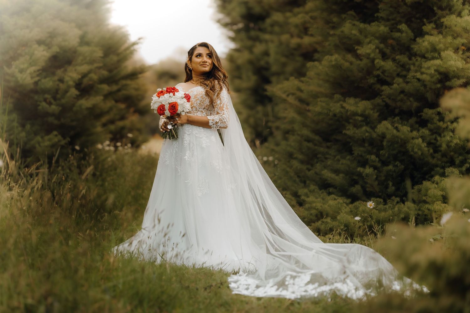 A bride in a flowing white gown with a long train holds a pink bouquet while standing in a grassy outdoor setting.