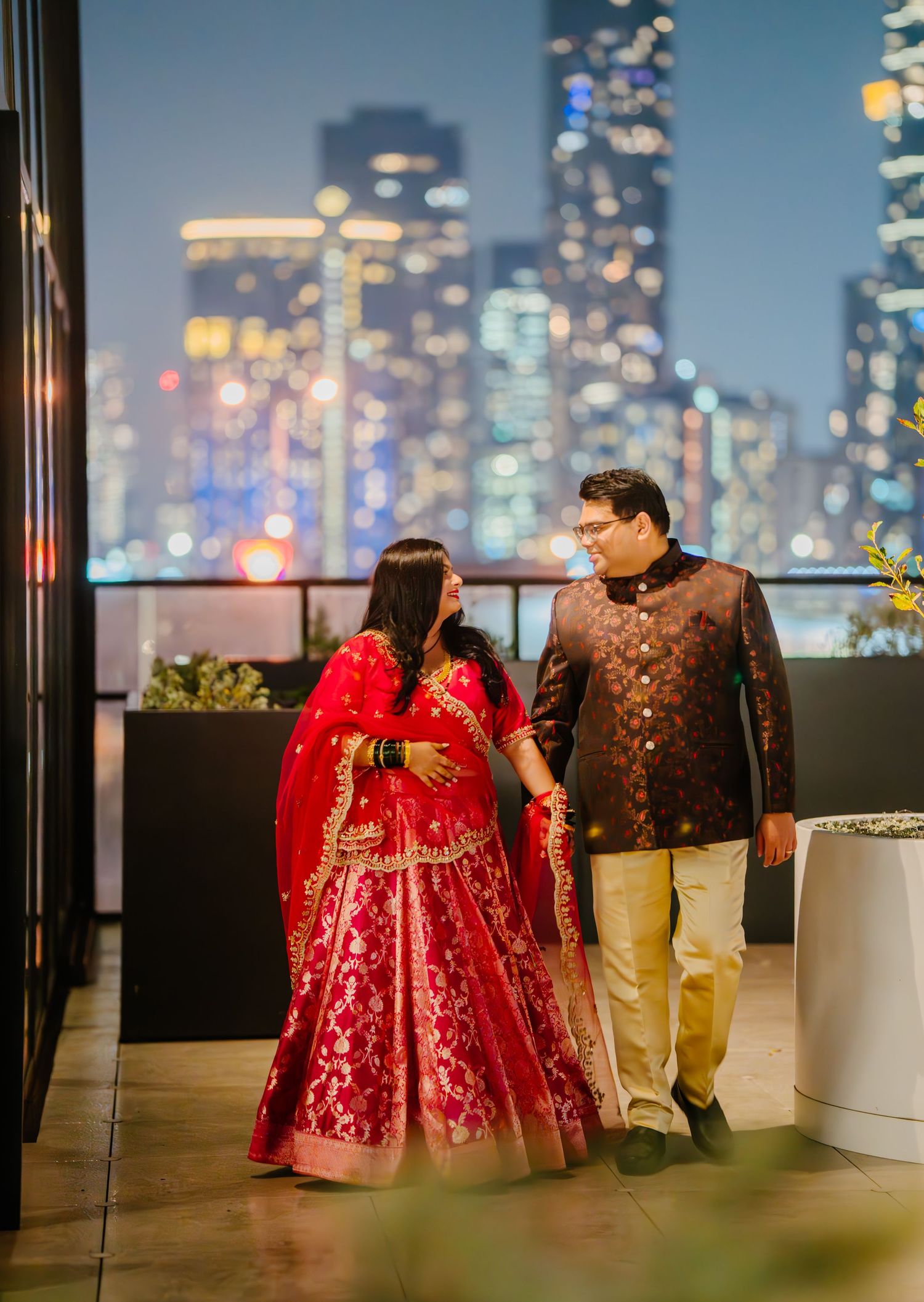 Couple in elegant red attire having a romantic moment against a glittering city skyline at night.