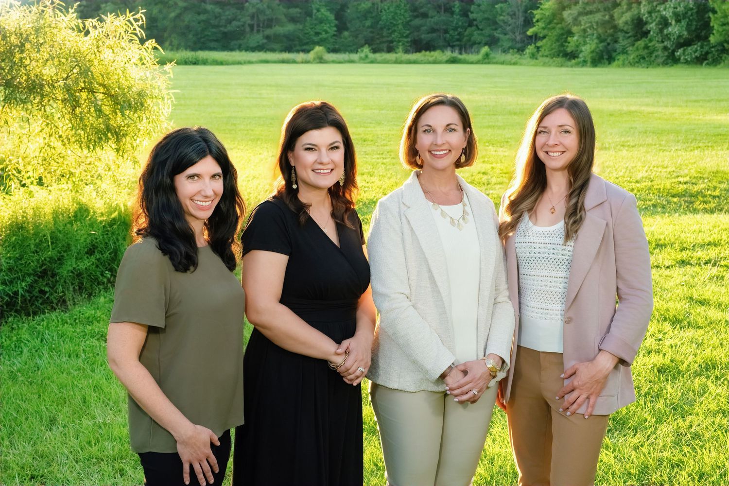 Four women pose for a professional photo together on a sunny day in a lush green outdoor setting.