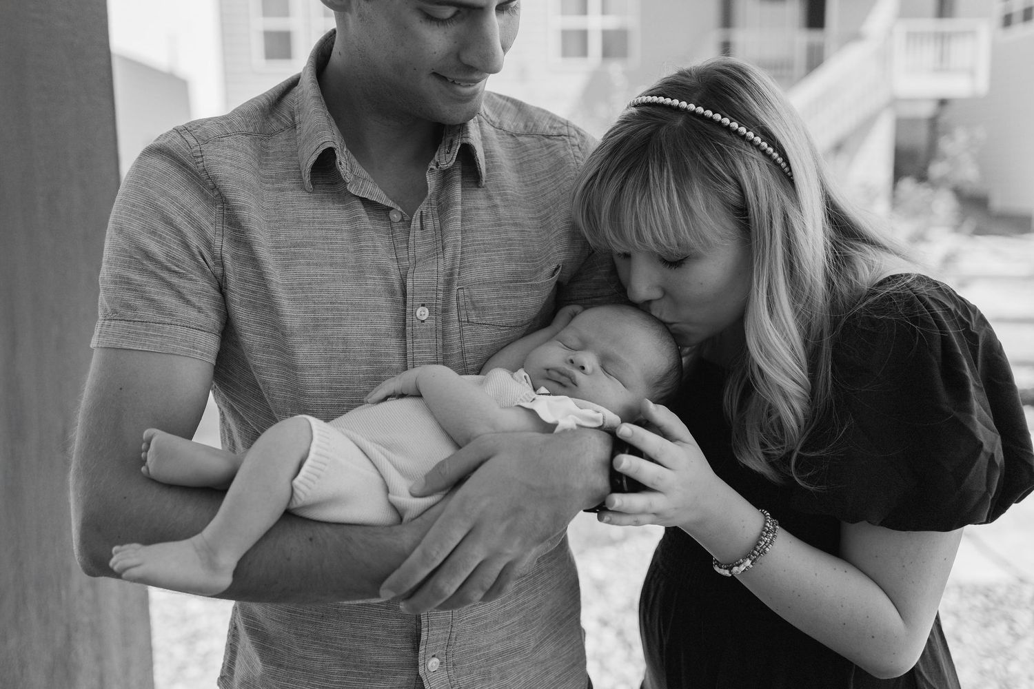 Black and white photo showing newborn lay in fathers arms.  Mother is tenderly kissing babys head.  Father is looking down and smiling