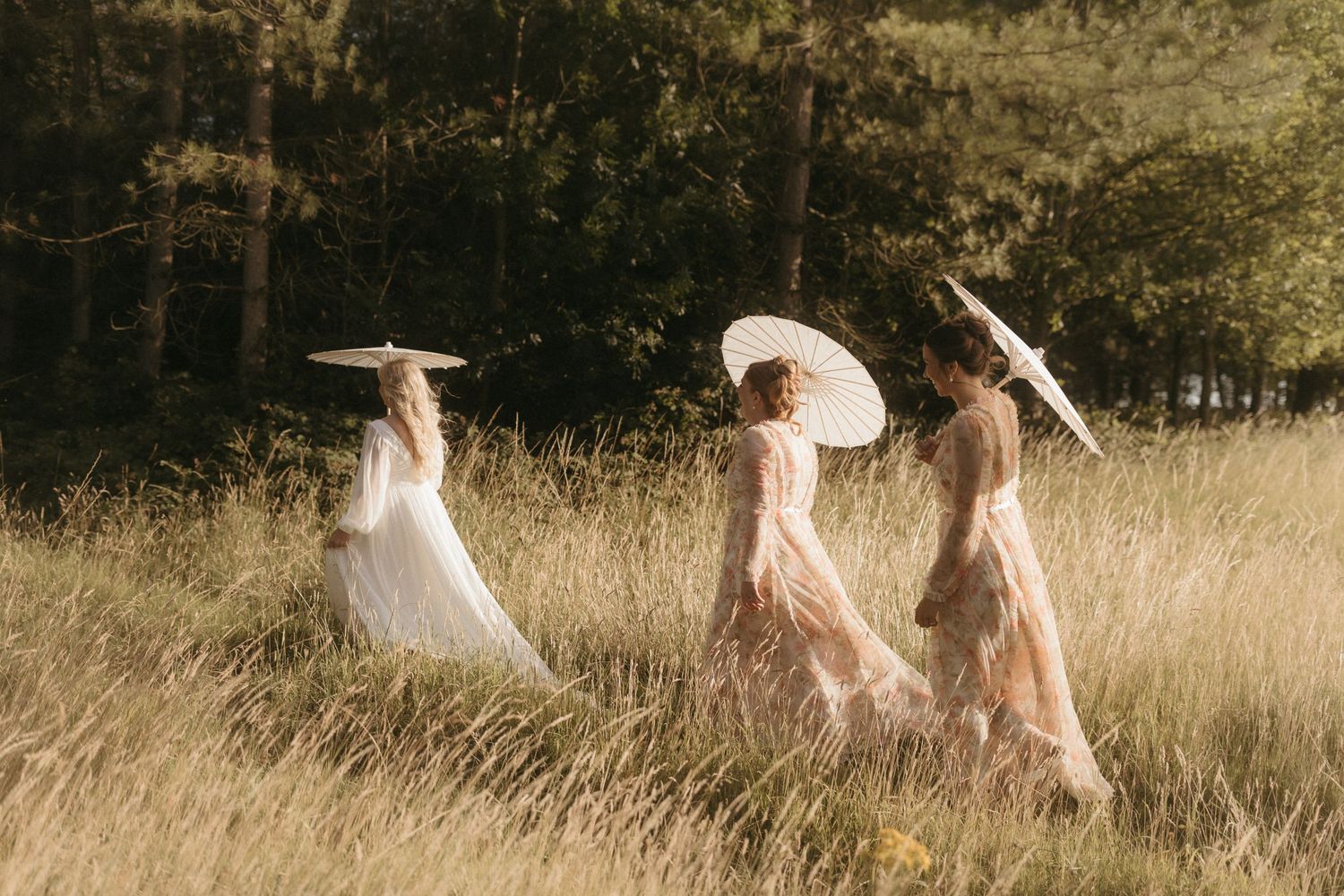 A group in dusty pink dresses holds white parasols and a flowing veil on a lush green lawn during a wedding ceremony.