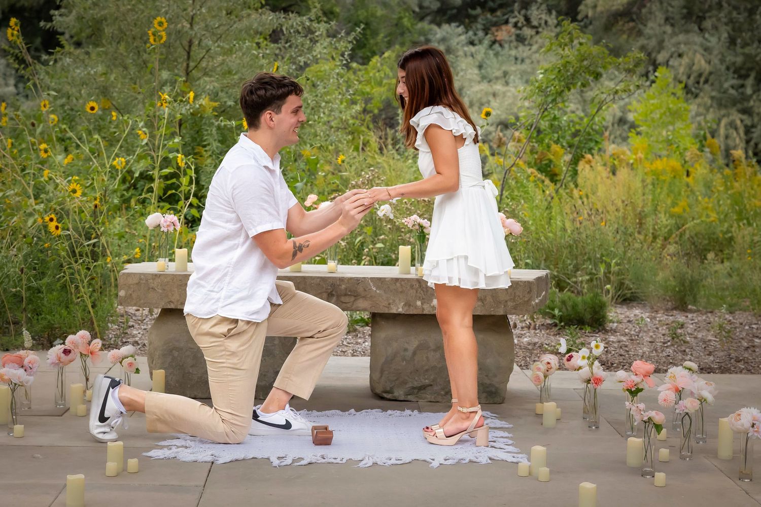 Man, on one knee, proposing to woman in park surrounded by candles and flowers.