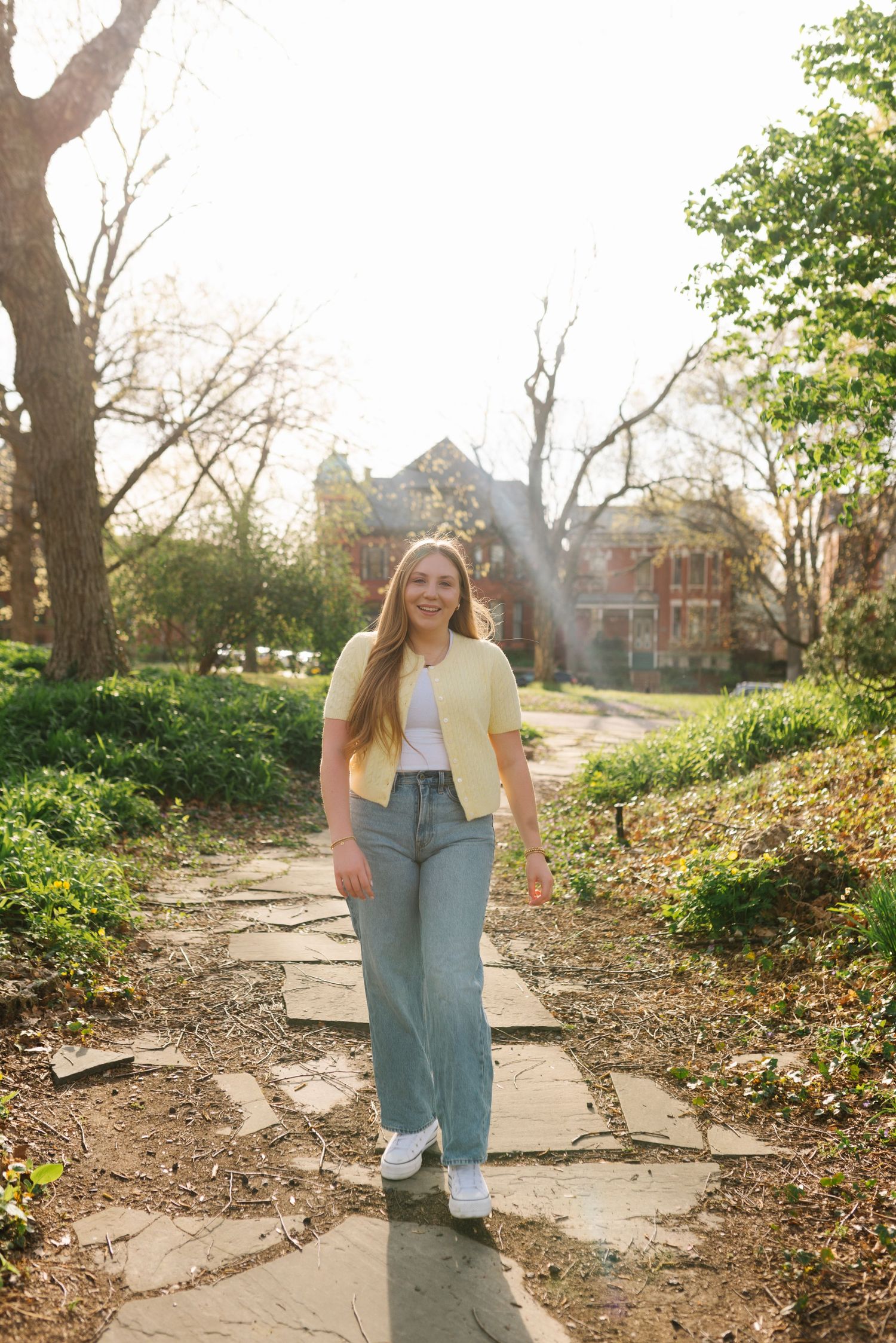 A person in a yellow cardigan and jeans walks along a stone path in a sunlit garden with trees and greenery.