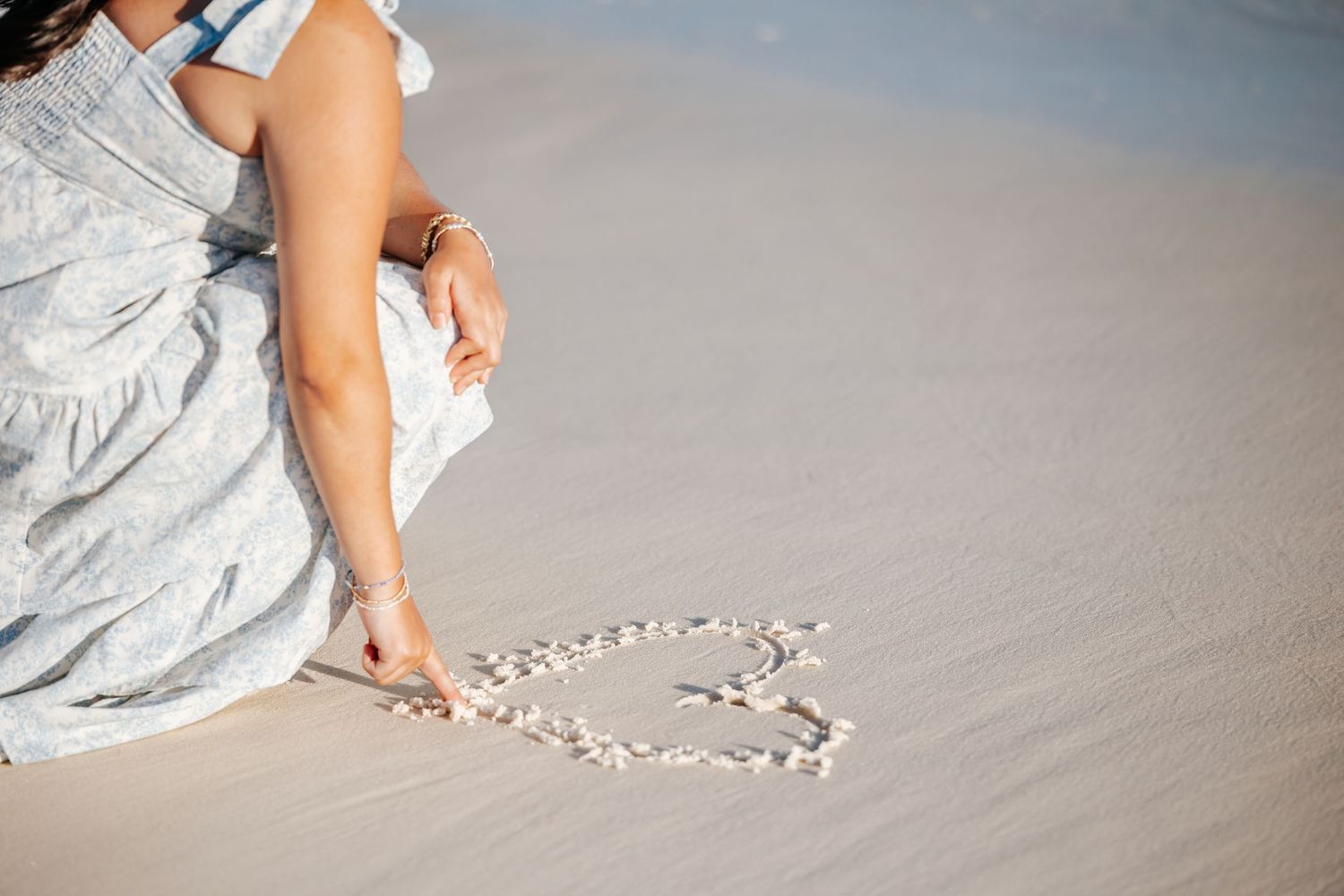 Hands arranging small white seashells into a heart shape on smooth wet sand at the beach.