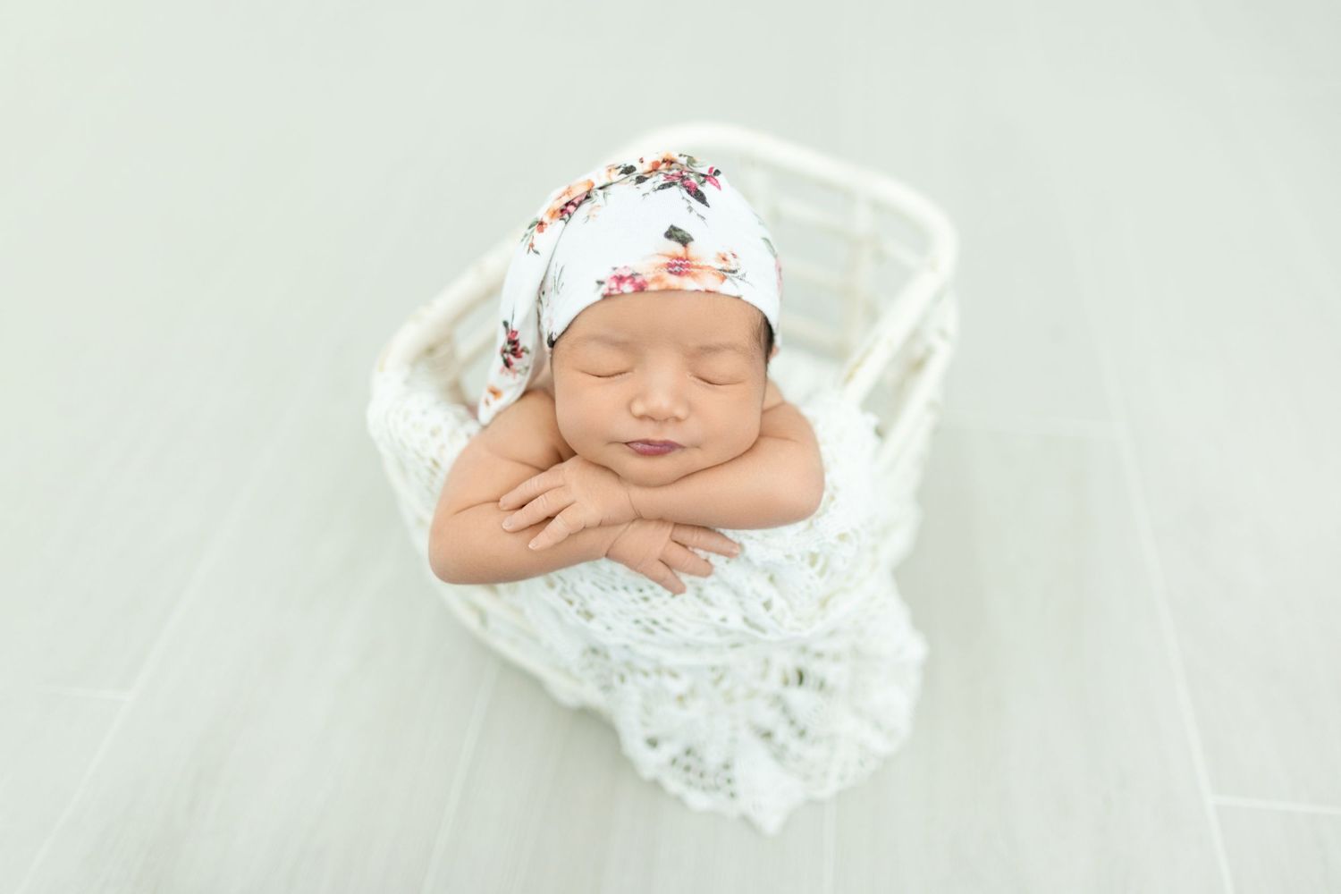 Newborn baby wearing white lace wrap and bonnet lies peacefully on white blanket during professional photo session.