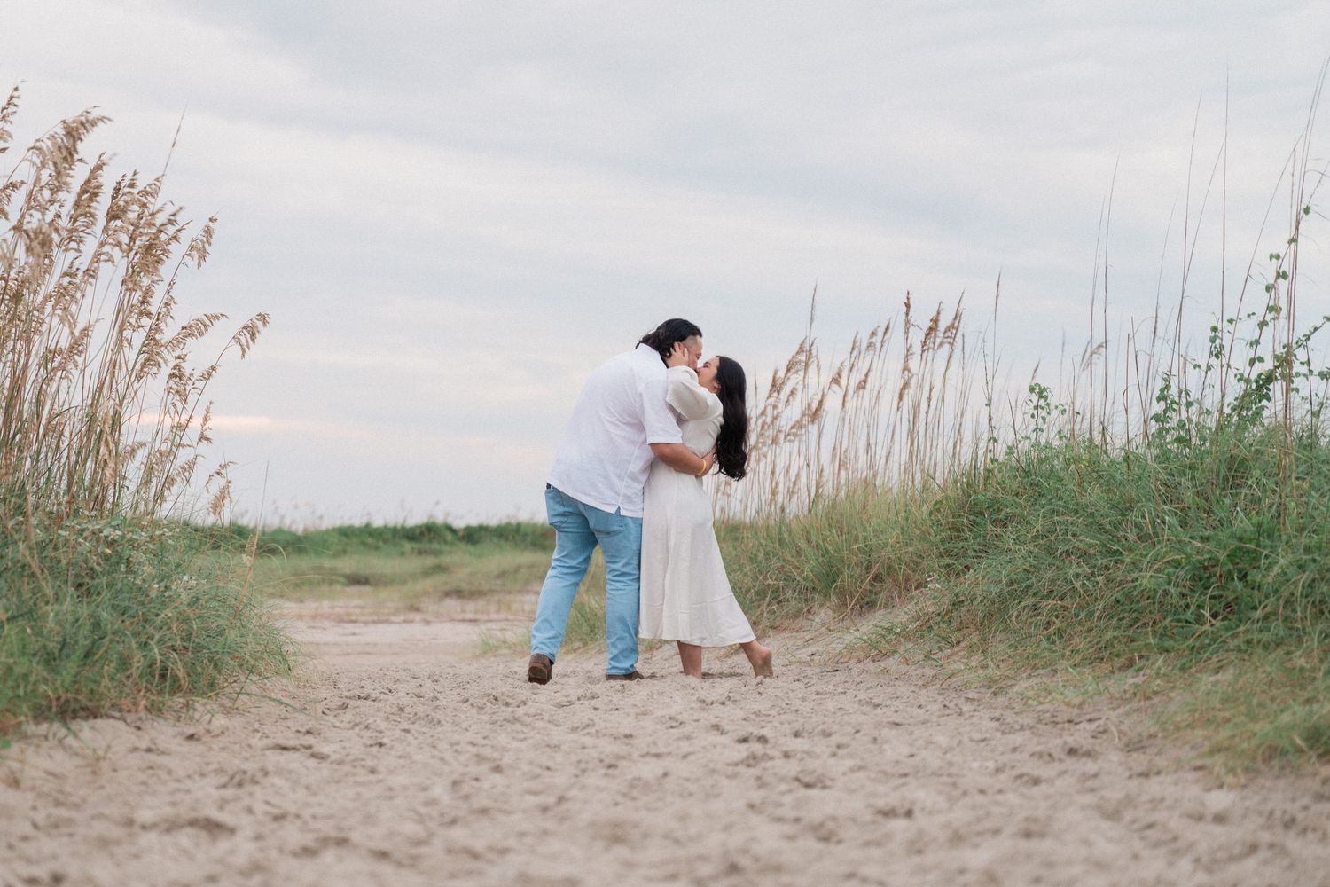 A couple walking along a sandy beach path through sea oats at sunset creating a romantic silhouette.