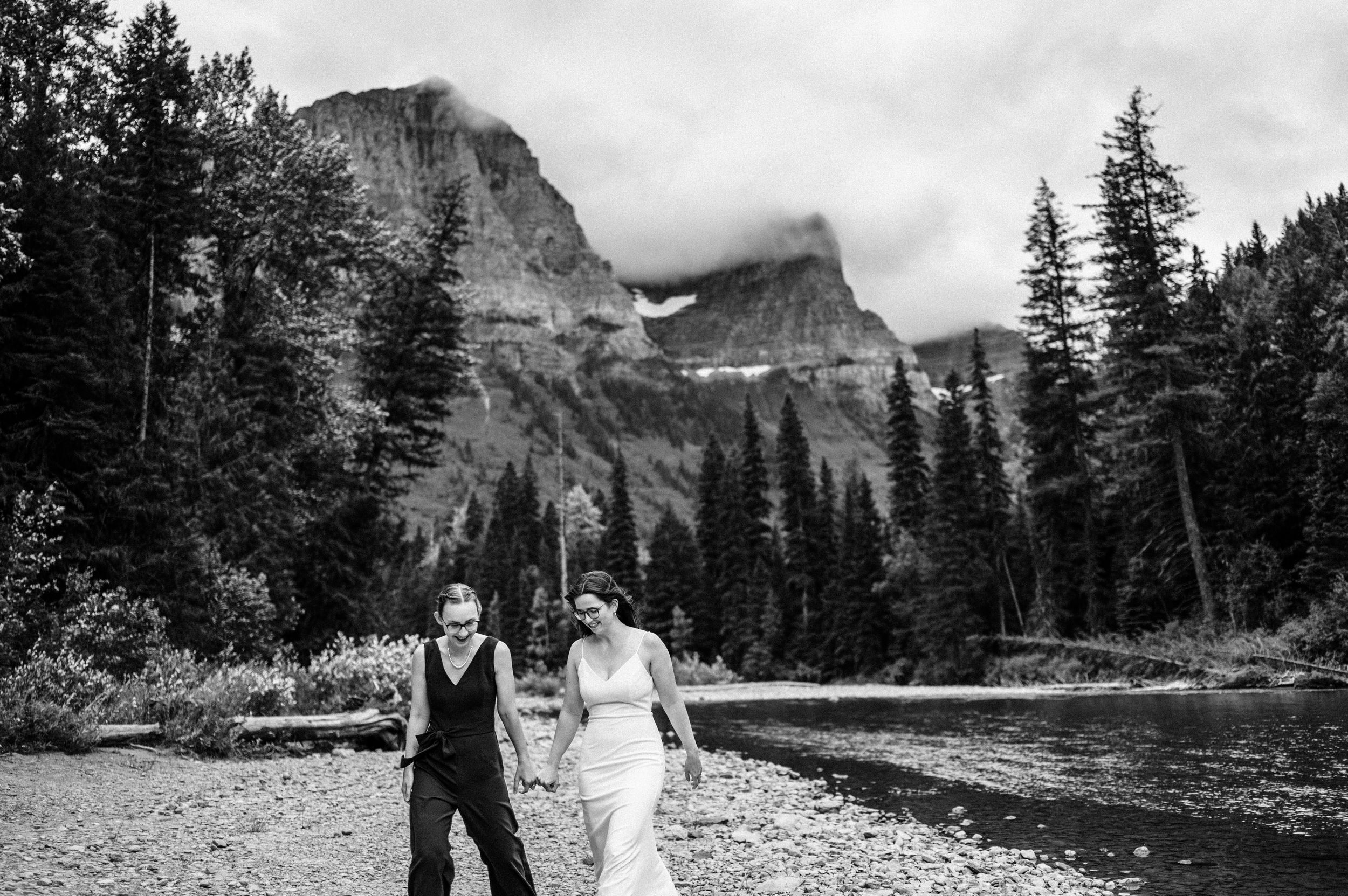 A couple walks along a river shore surrounded by evergreen trees and dramatic rocky peaks in black and white. Avalanche Amphitheater Wedding