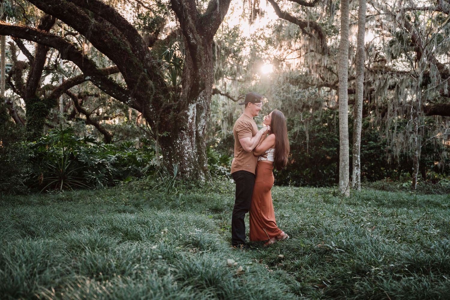 A romantic couple embraces under a majestic oak tree draped with Spanish moss at sunset in a lush green field.