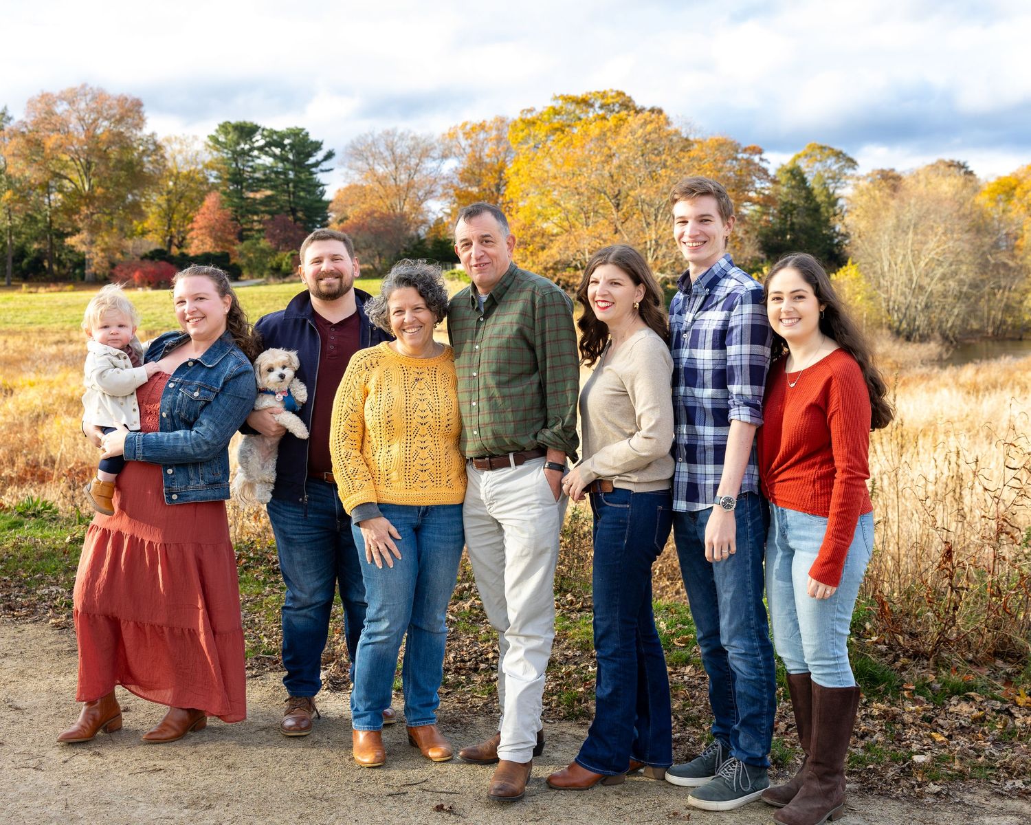A A multi-generational family poses casual fall attire pose together outdoors with autumn foliage and golden fields in the background.