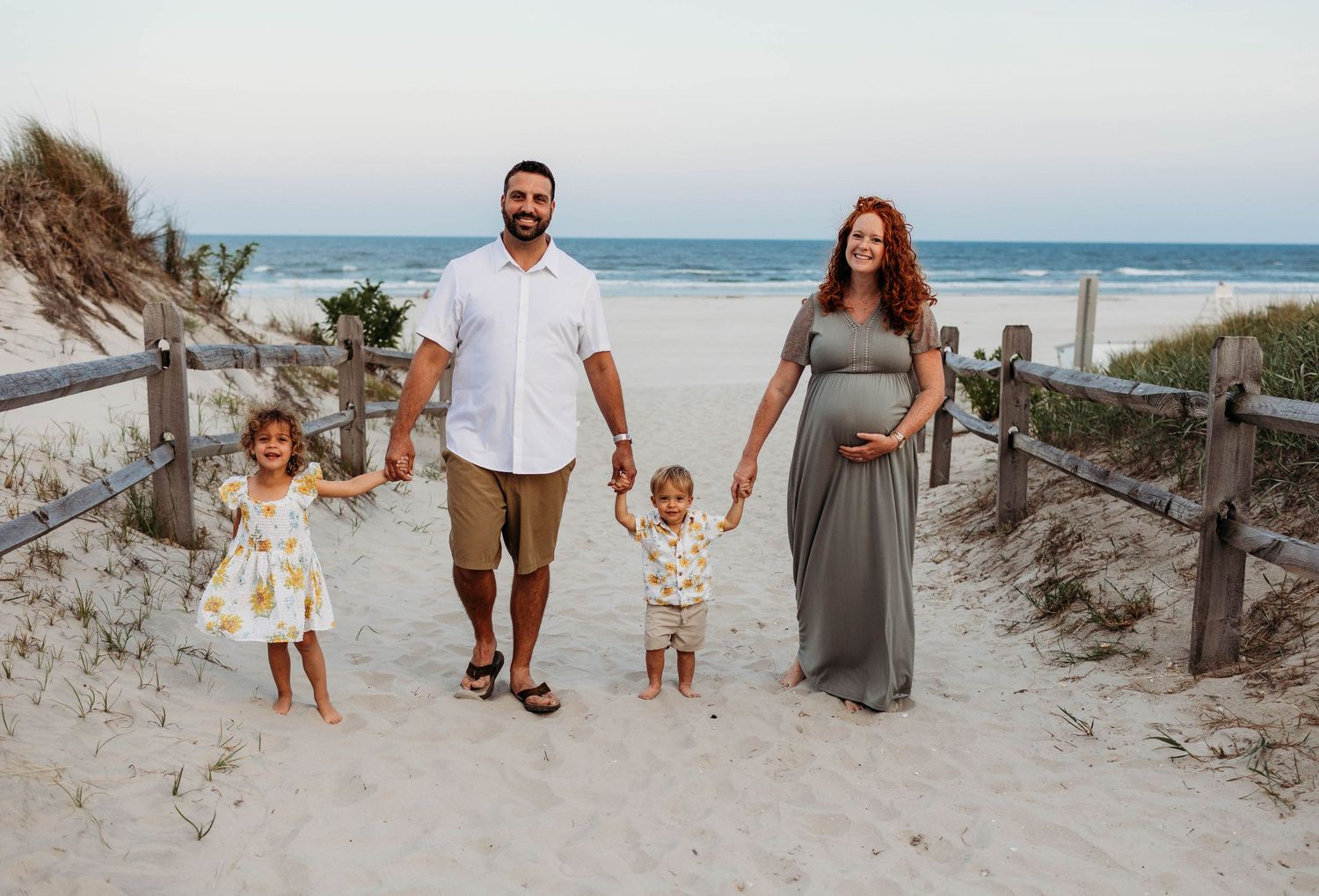 A family walks together on a sunny beach boardwalk near sand dunes and ocean waves during sunset.