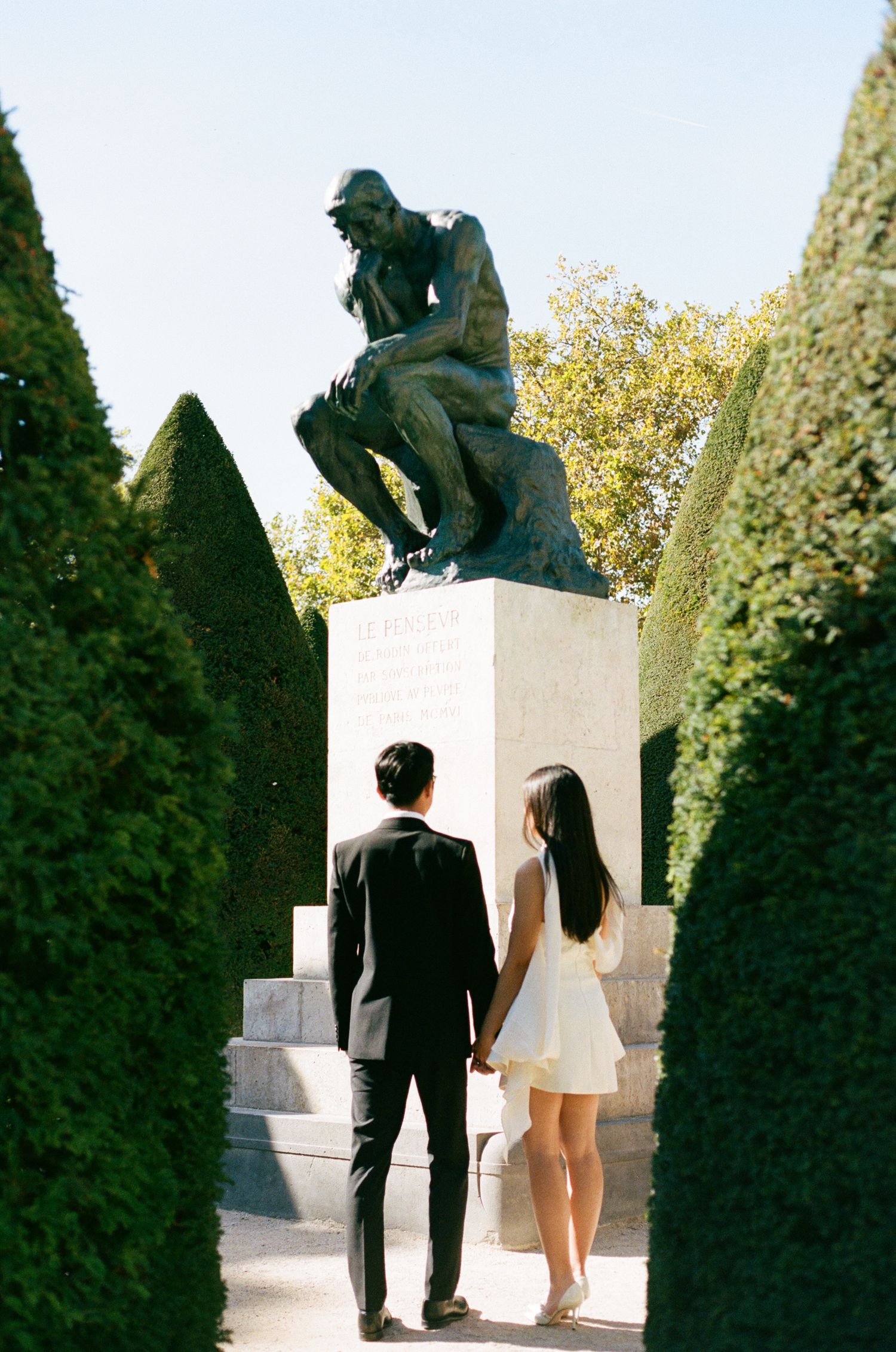 A couple admires Rodin's The Thinker sculpture in a garden with manicured hedges on a sunny day.