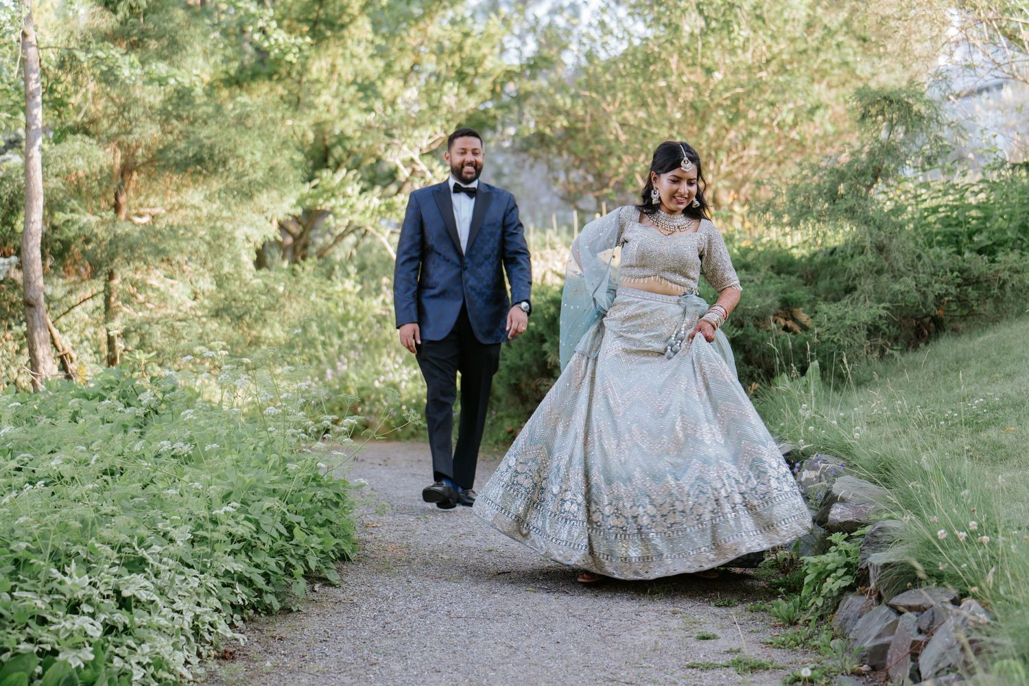 A couple in traditional South Asian wedding attire walks along a garden path surrounded by greenery.
