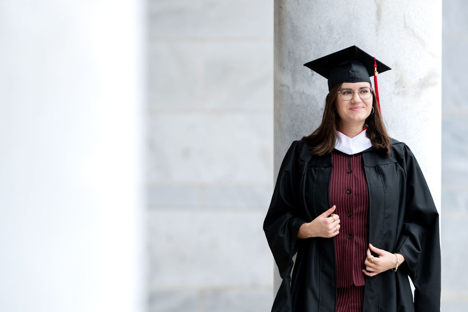 Young woman in black graduation cap and gown smiling between marble columns.