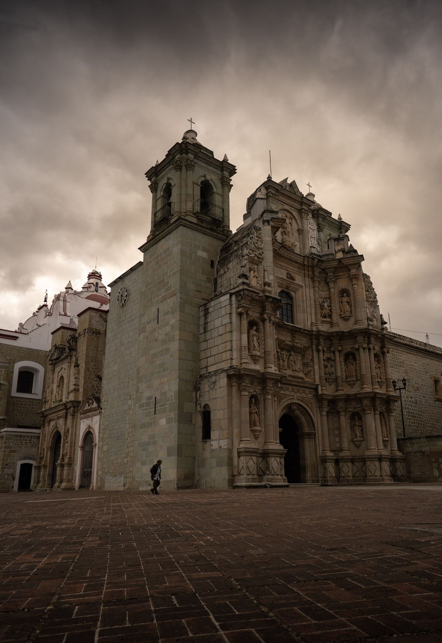 Historic baroque church facade in Oaxaca, Mexico, with dramatic cloudy sky and stone courtyard.