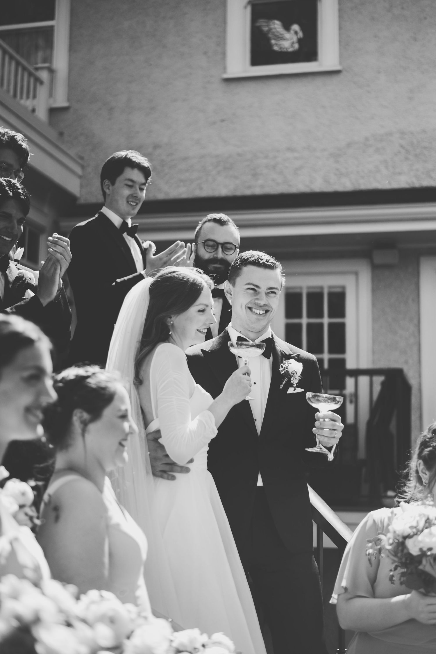 A black and white photo of a wedding celebration with a bride in white dress and guests sharing joyful moments outdoors.
