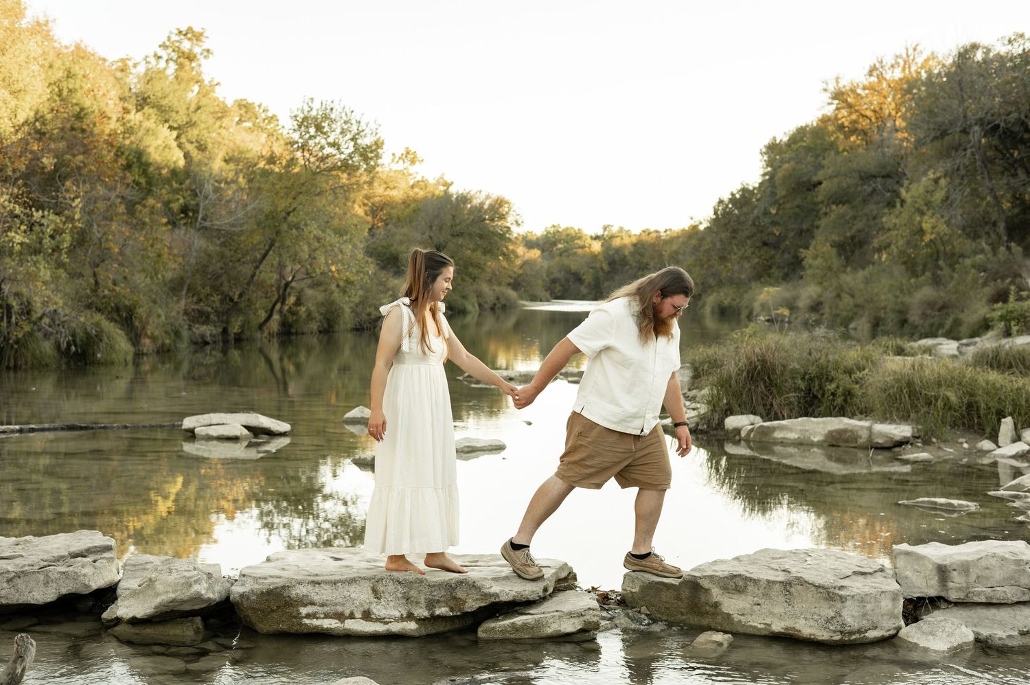 Romantic sunset photoshoot on a rocky riverbank with golden light filtering through autumn trees.