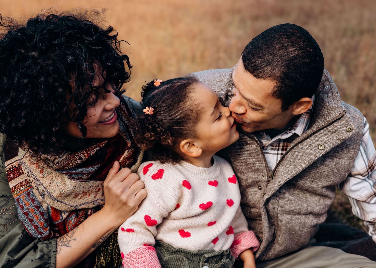 A joyful family moment outdoors with parents embracing and kissing their young child wearing a heart-patterned sweater.