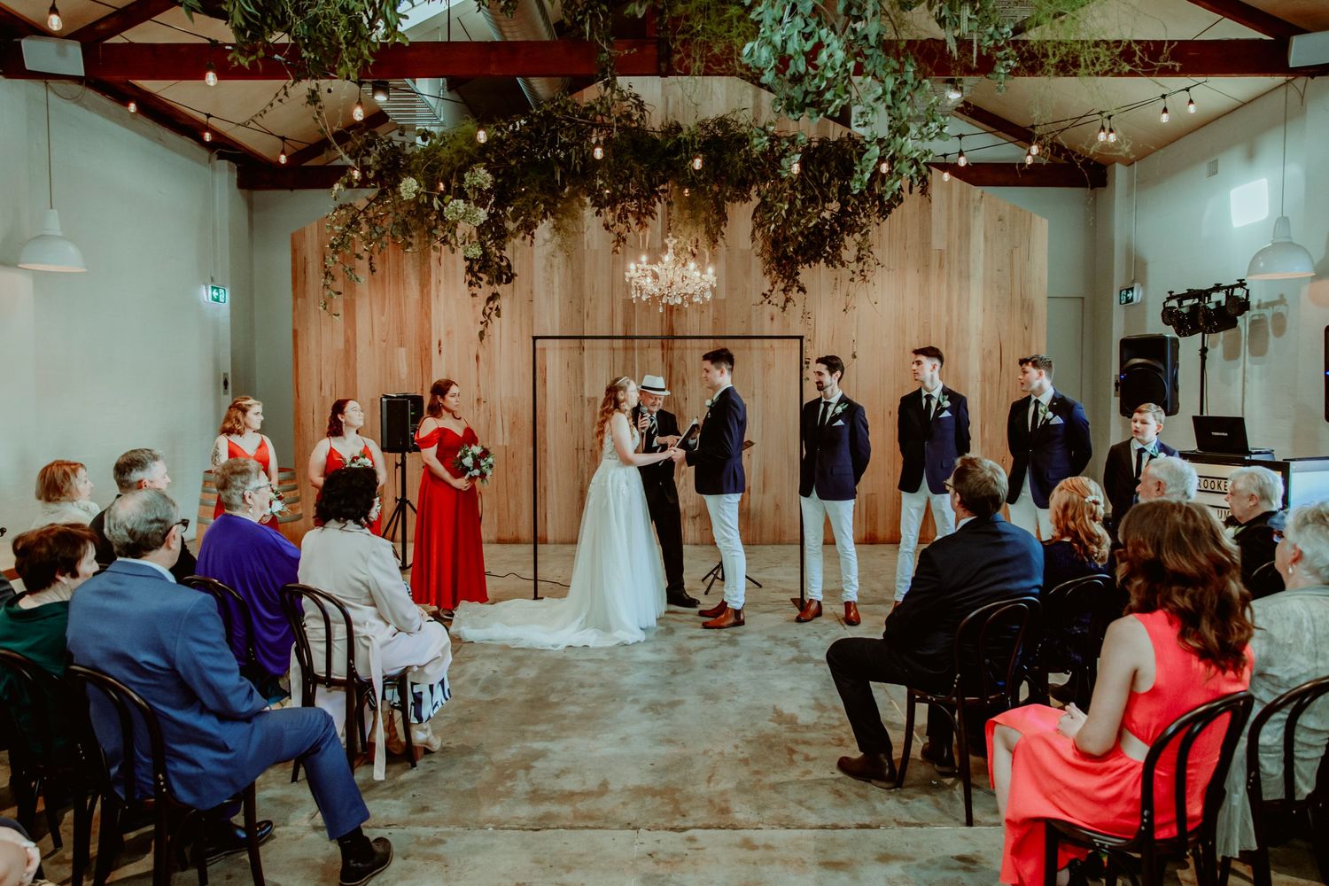 Indoor wedding ceremony taking place in an elegant venue with chandelier and greenery decor hanging from exposed beams.