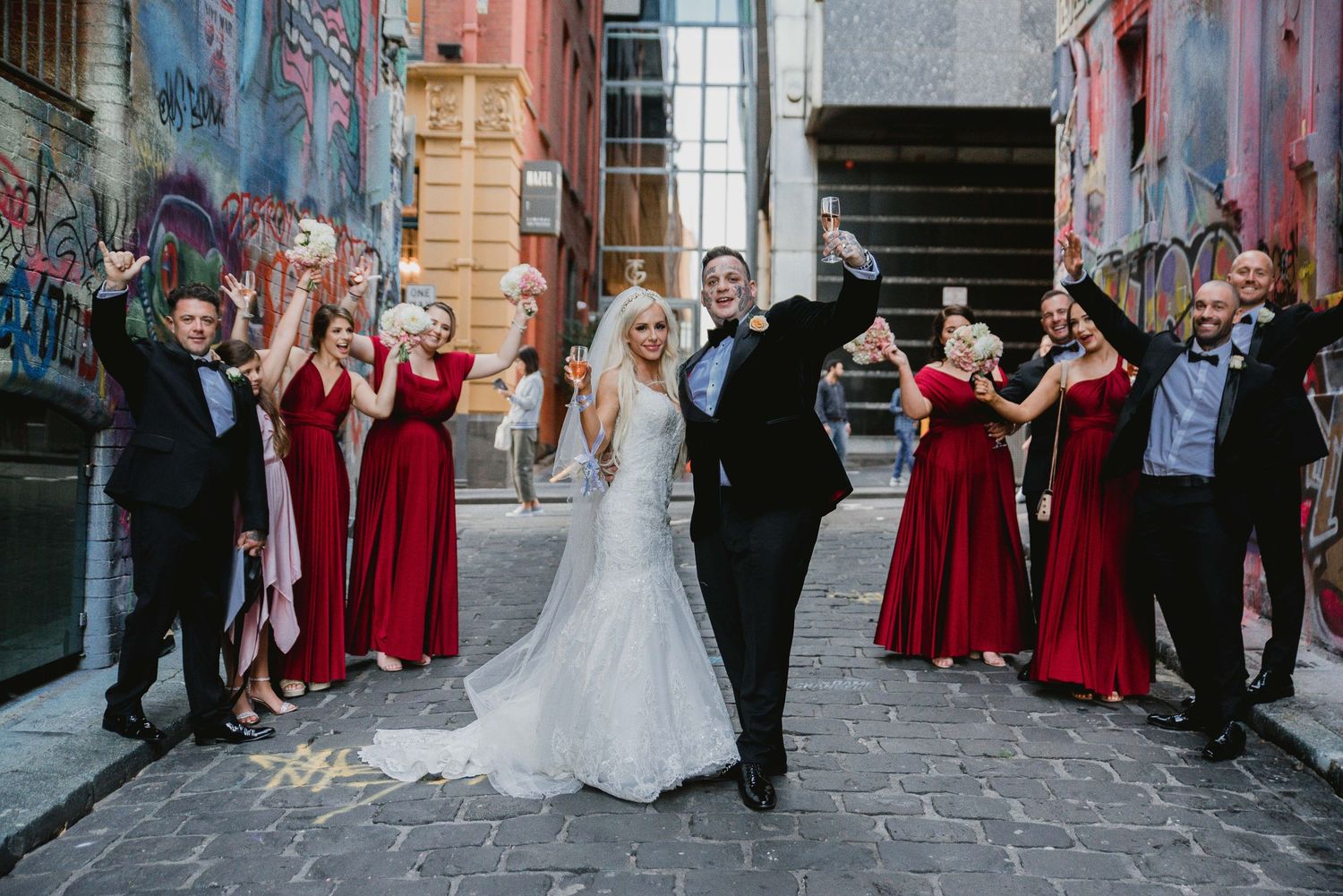 Wedding party celebrates in urban alley with bridesmaids in red dresses and colorful street art backdrop.