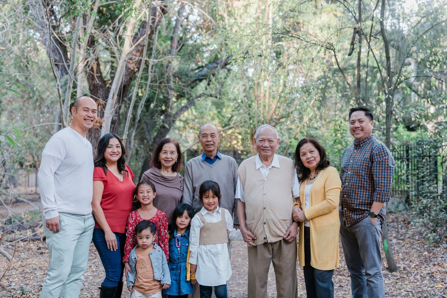 A multi-generational family poses together outdoors on a wooded path wearing coordinated casual attire.