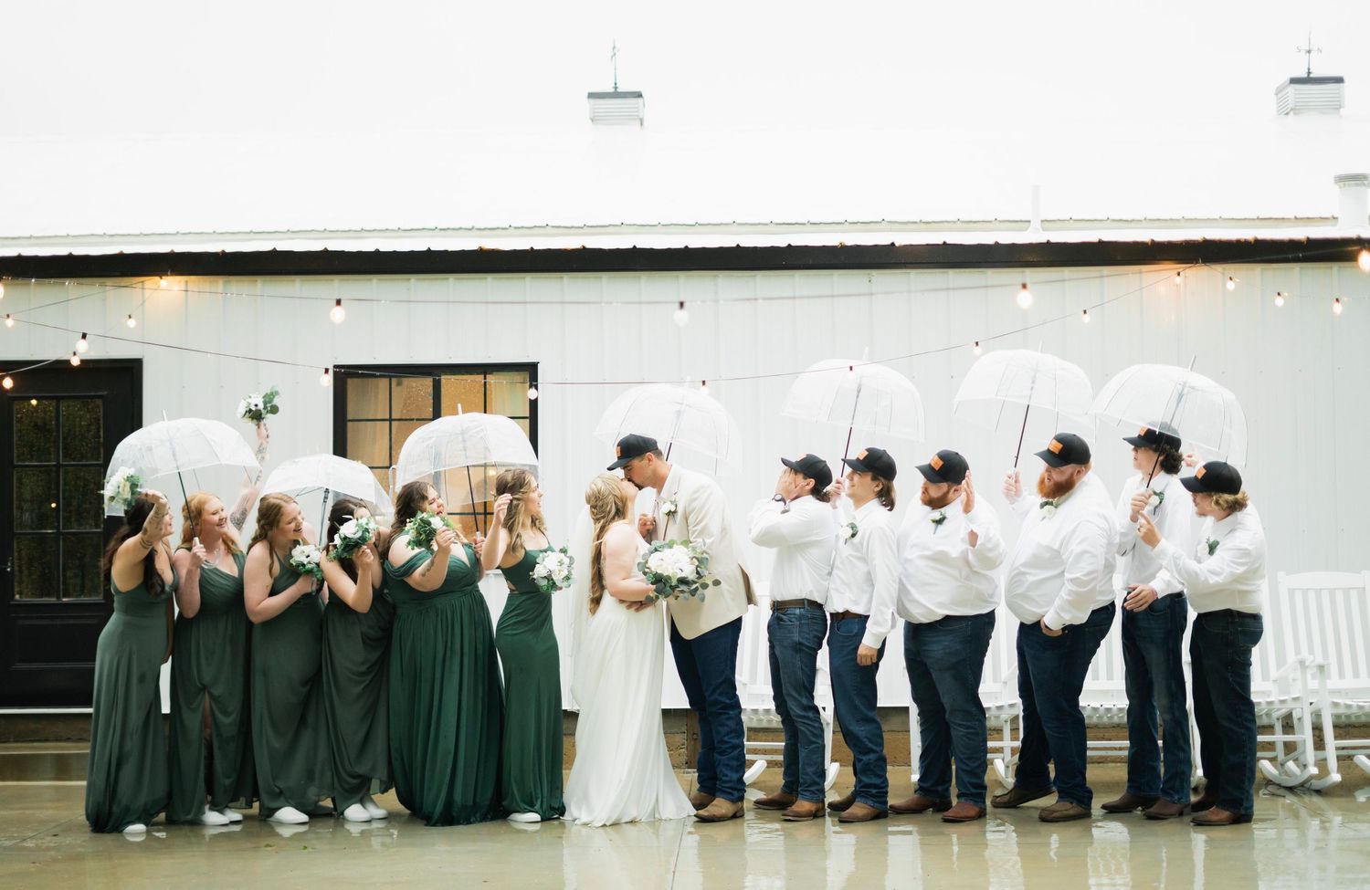 A wedding party holds white umbrellas in the rain outside a white barn venue, bridesmaids in green.