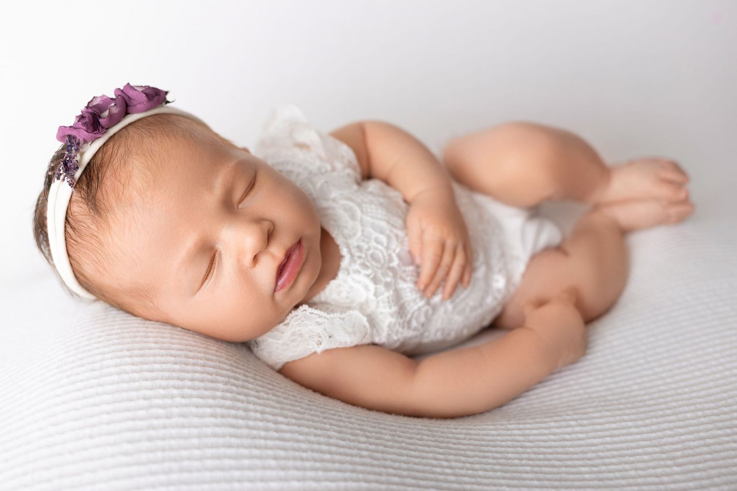 A sleeping newborn wearing white lace and a purple floral headband rests on a textured white surface.