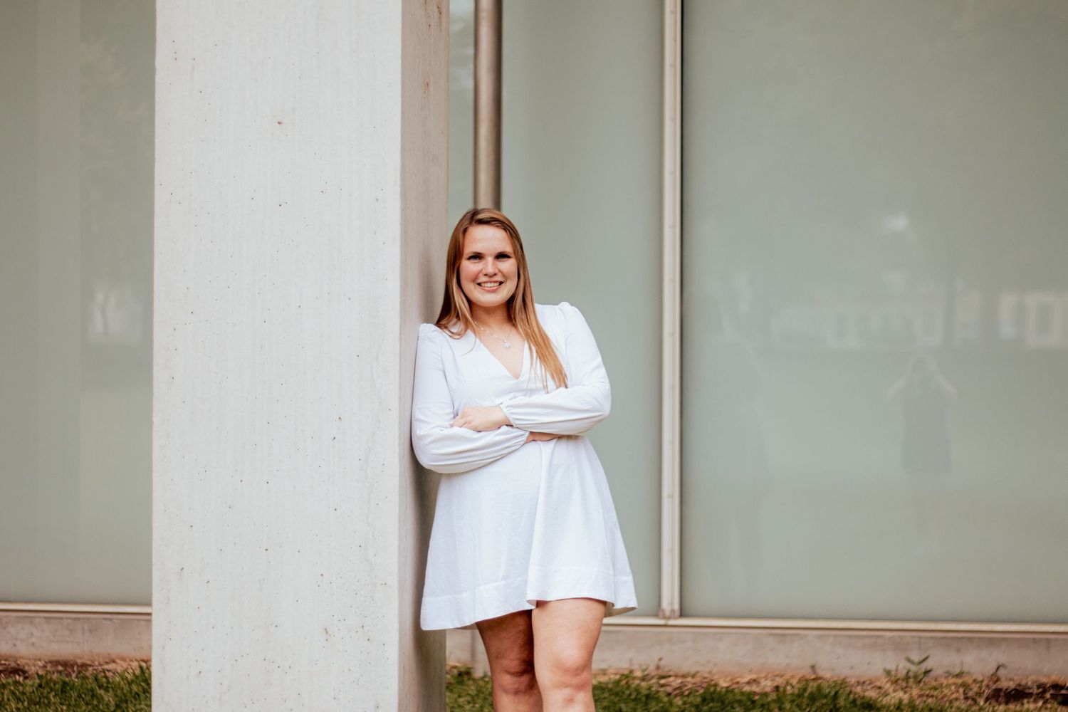 Confident person in a white flowing dress poses with arms crossed against a neutral wall outdoors.