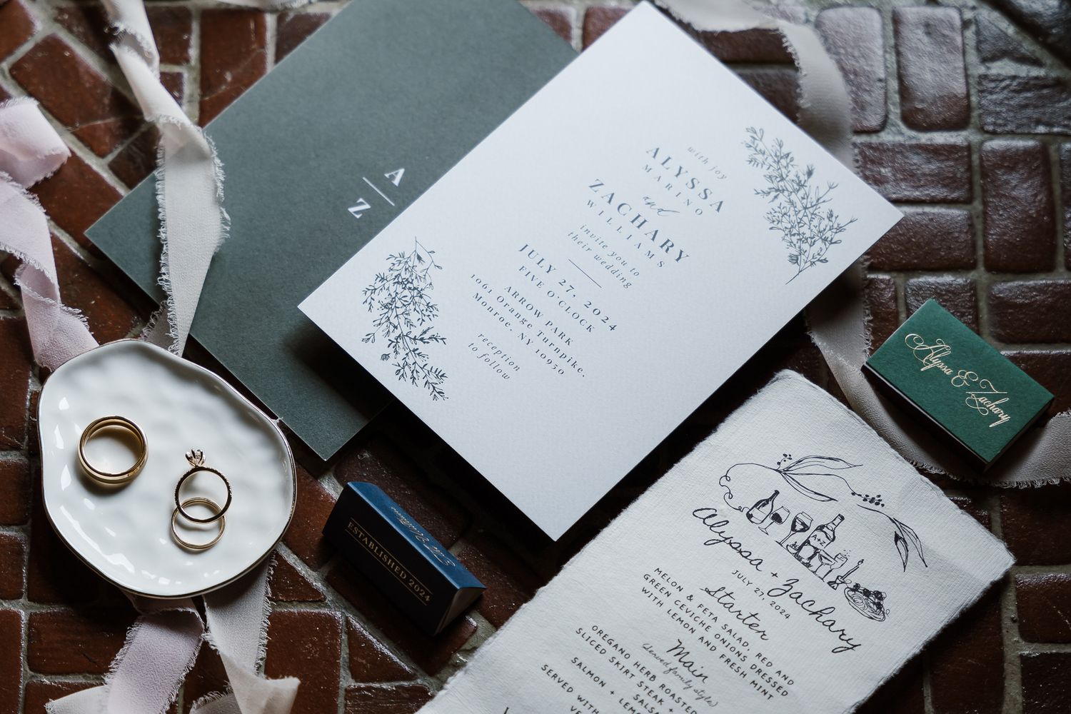 Wedding invitations and rings displayed on marble with sage green accents and floral designs.