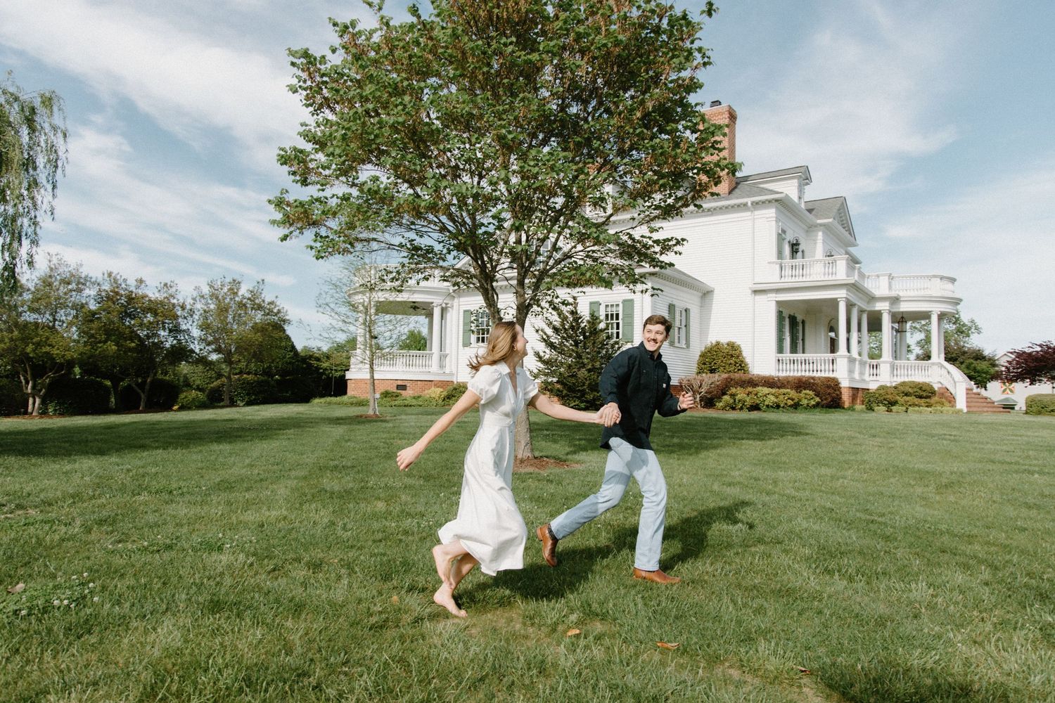 Two people dance playfully on a green lawn in front of a white historic Victorian-style house on a sunny day.