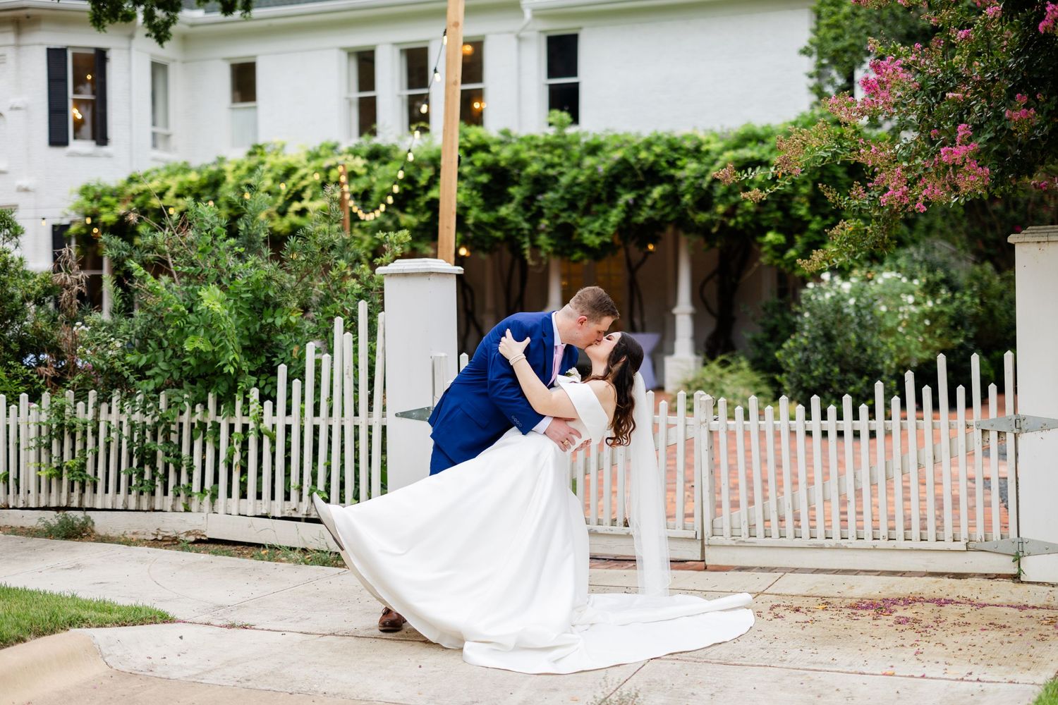 Romantic wedding couple sharing a dip pose in front of a white picket fence and garden.