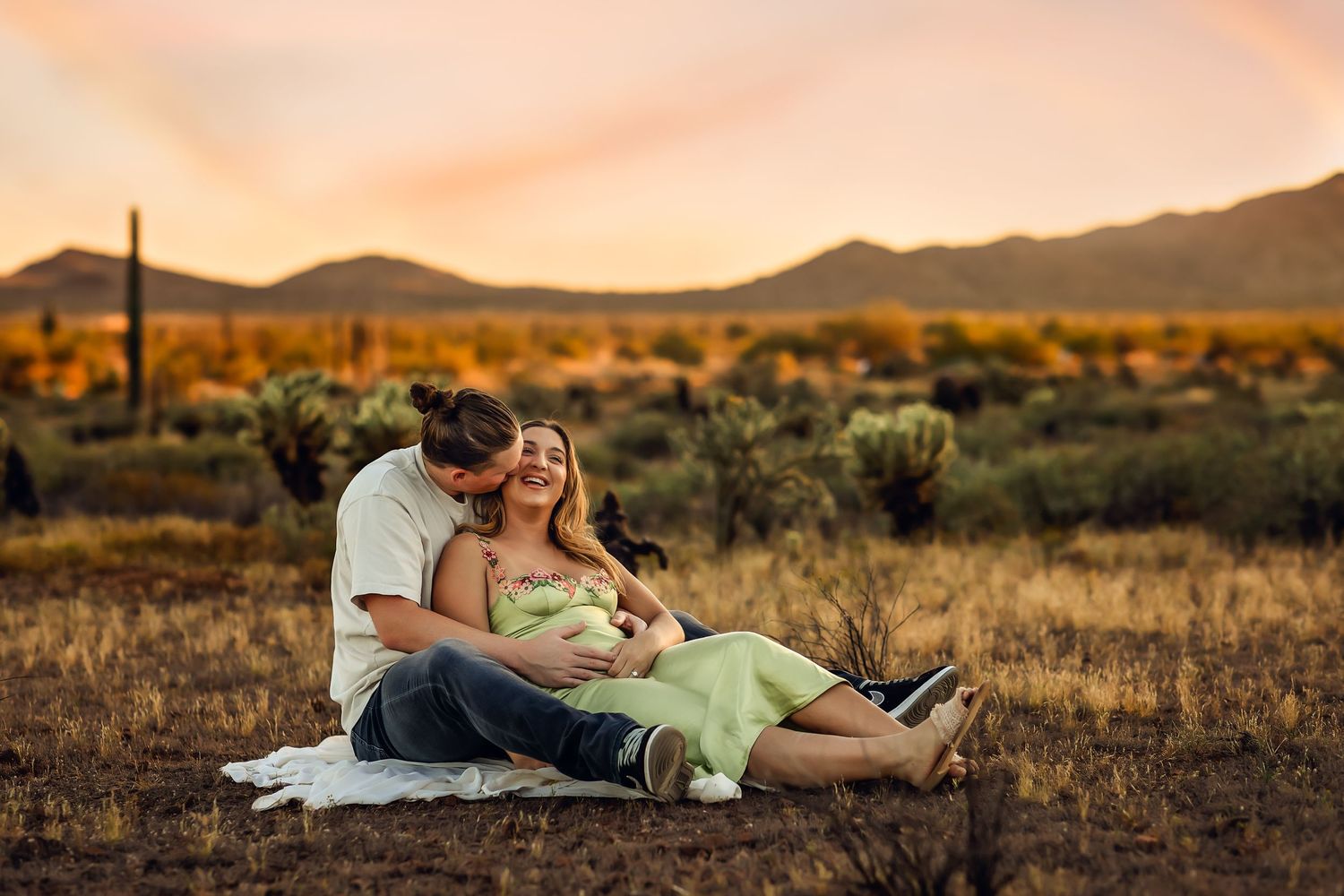 Couple sits together on blanket in desert landscape during golden sunset with cacti and mountains in background.