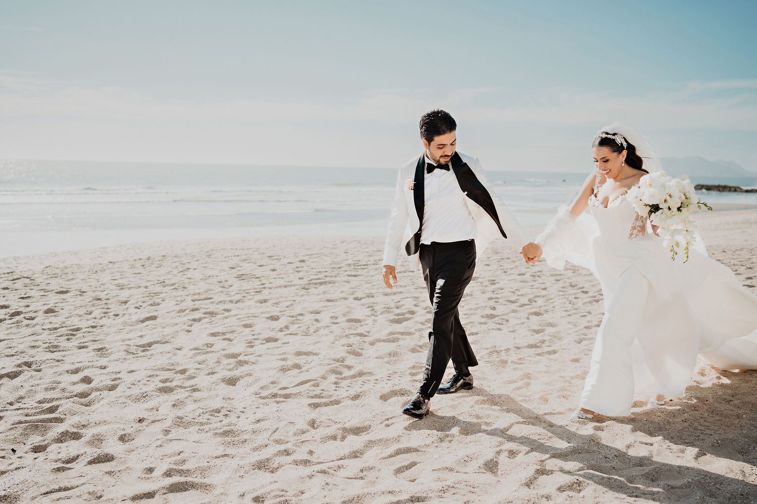 A couple walks together on a sunny beach, with waves and blue sky in the background.