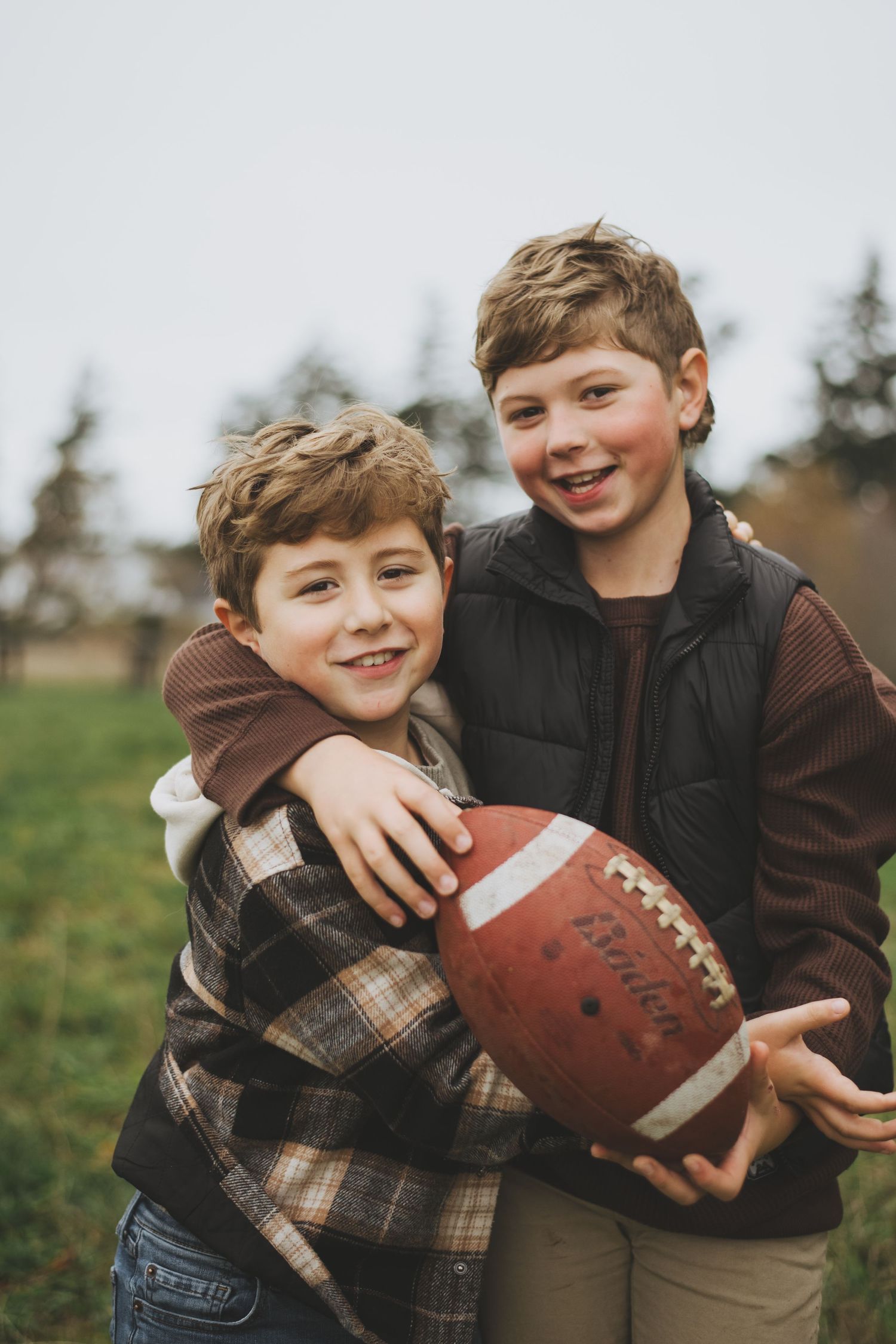 Two young brothers in casual fall clothing share a football while playing outdoors in a grassy field.