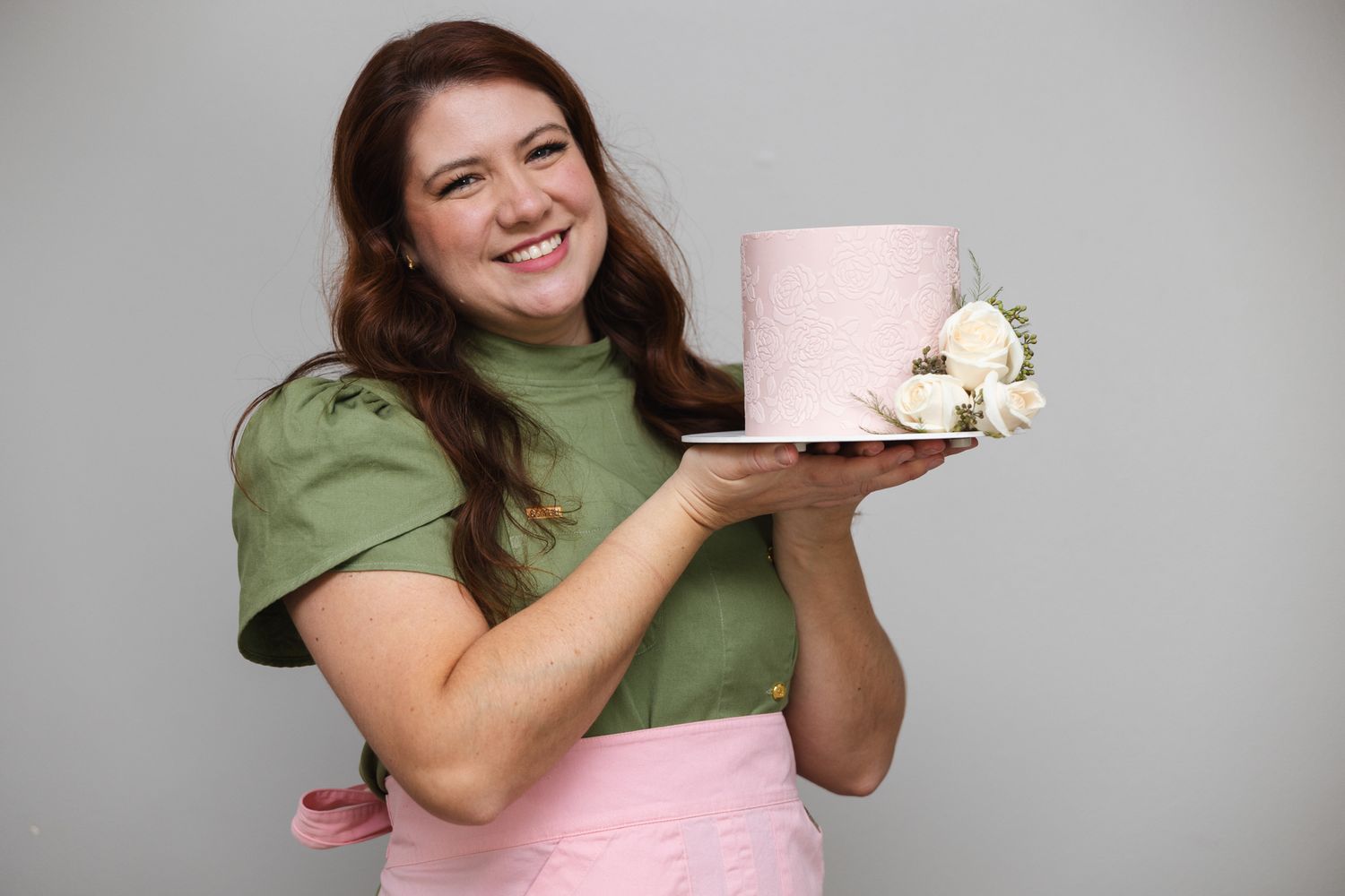 Baker wearing green top and pink apron holds pink cake on plate with white flower decoration.