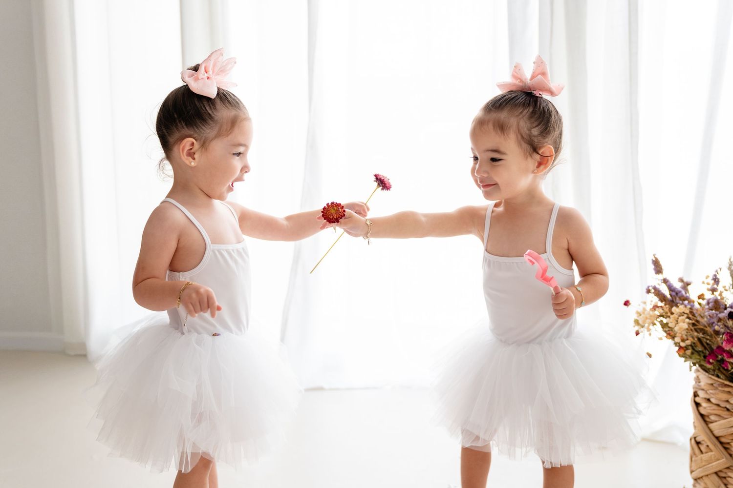 Two young ballerinas in white tutus and hairbows share a red flower while dancing in a bright sunlit room.