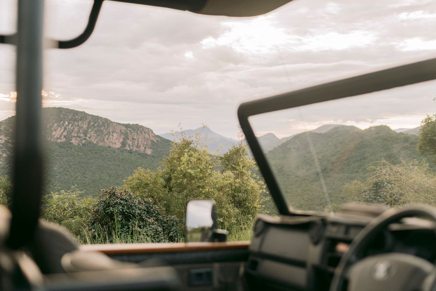 View from inside a safari vehicle overlooking lush green mountains under a cloudy sky.