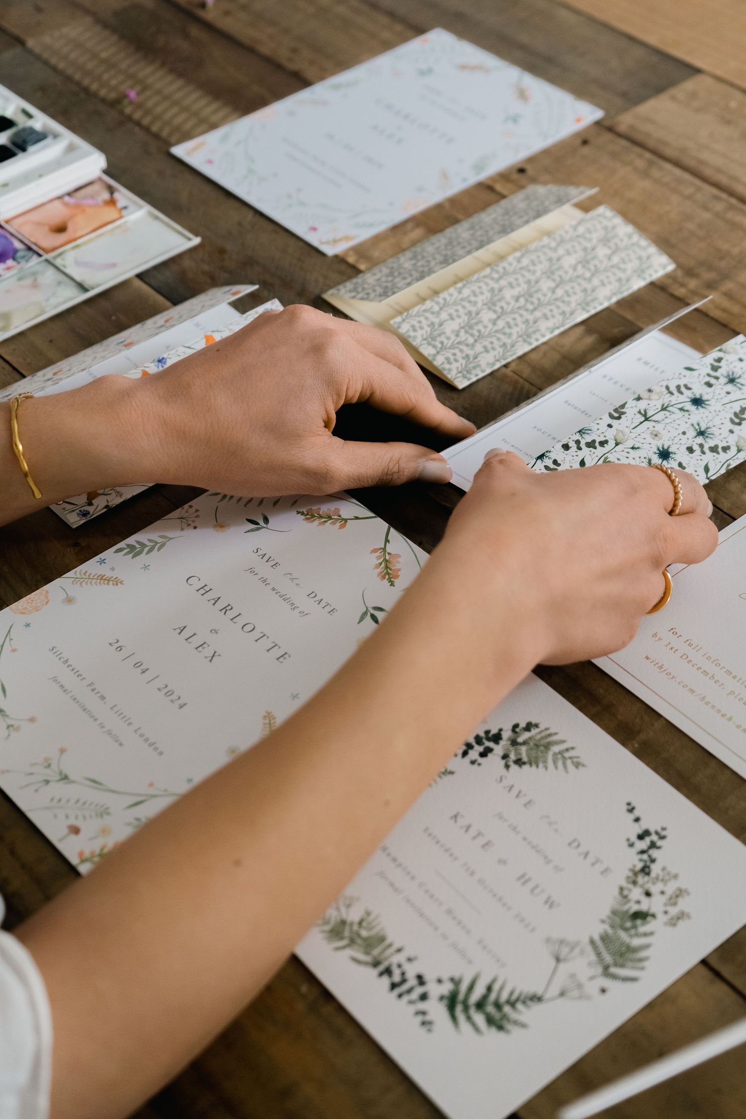 Hands arranging wedding invitations with floral designs on wooden table.
