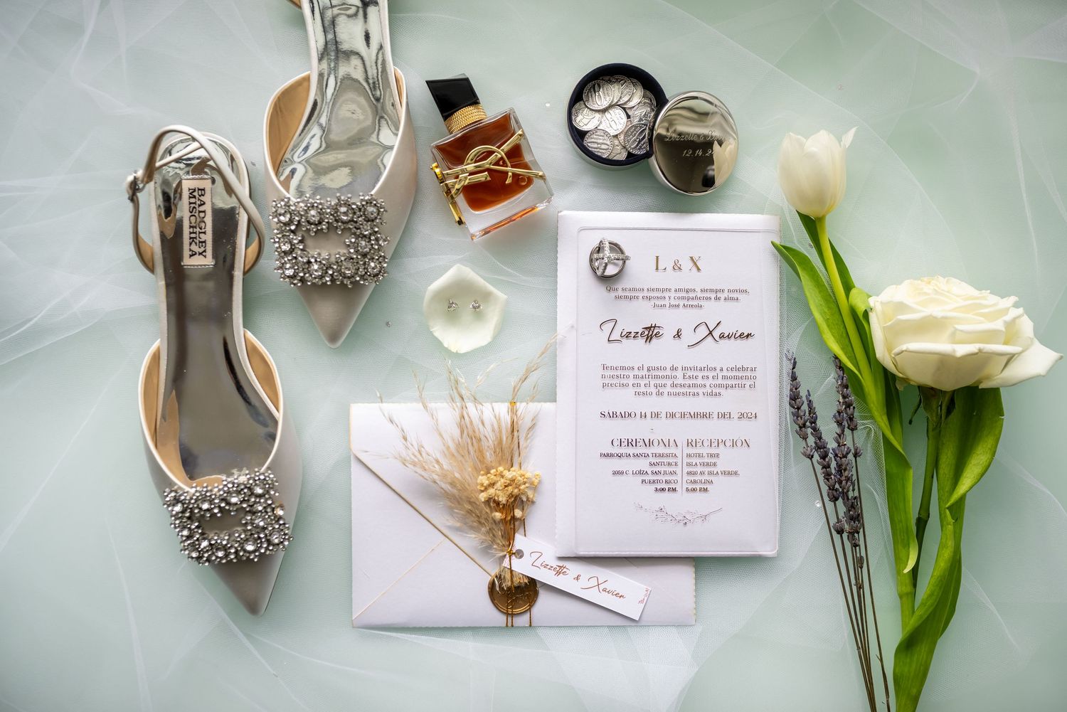 Wedding accessories including silver shoes, invitations, and white roses arranged on a light blue surface.