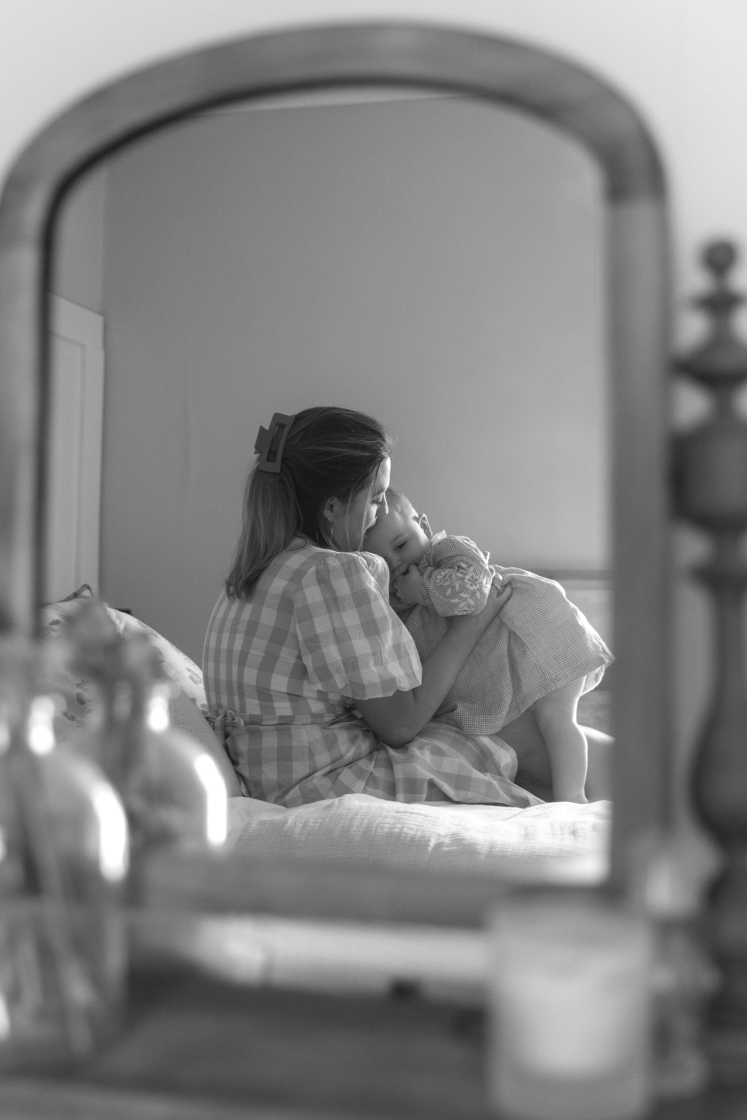 A tender black and white reflection in a mirror shows a parent holding and caring for their baby in a bedroom.