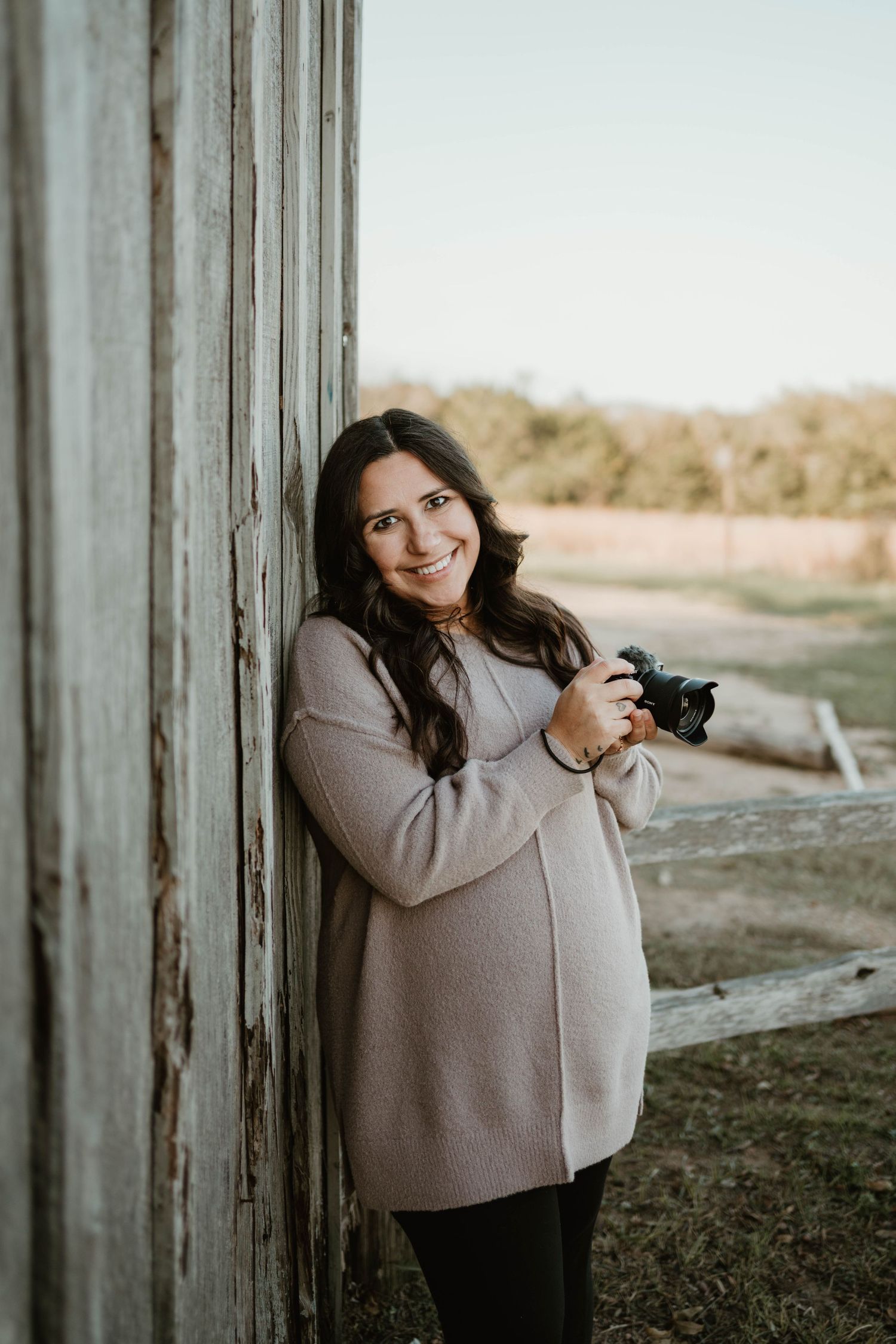 A photographer wearing a tan sweater holds a camera while standing next to a rustic barn wall at sunset.