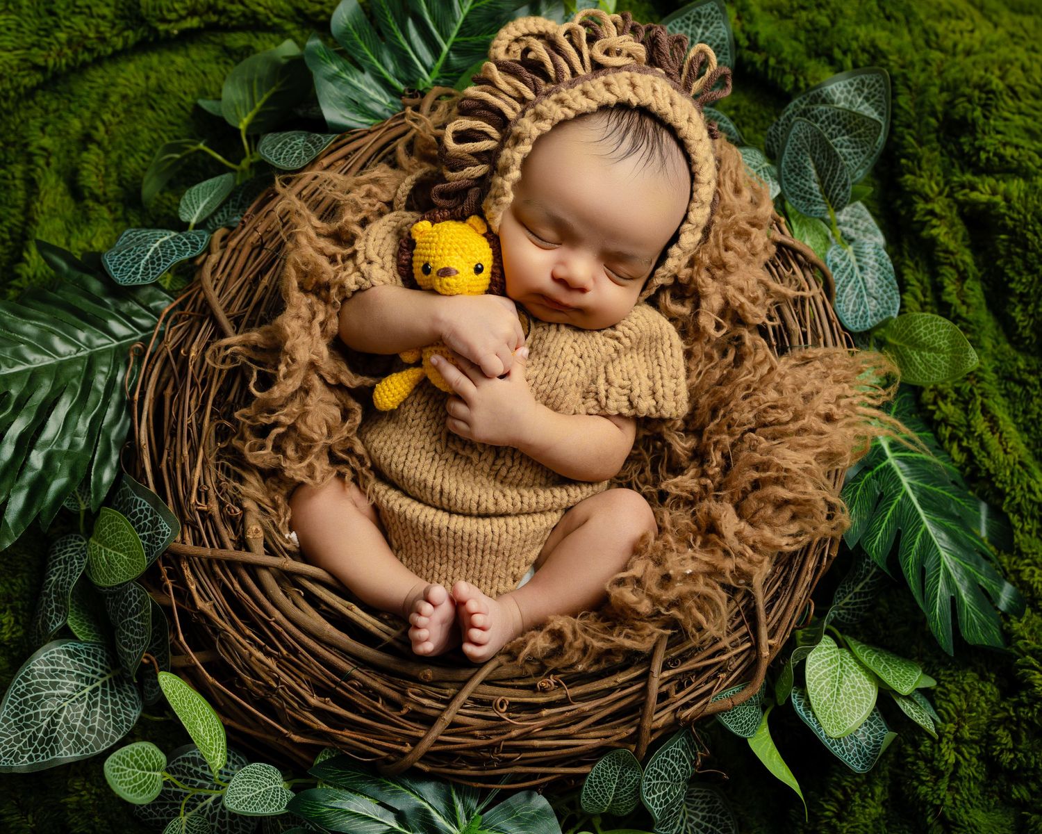 A sleeping baby in a brown knit outfit cuddles with a small yellow teddy bear in a nest-like basket surrounded by green leaves.