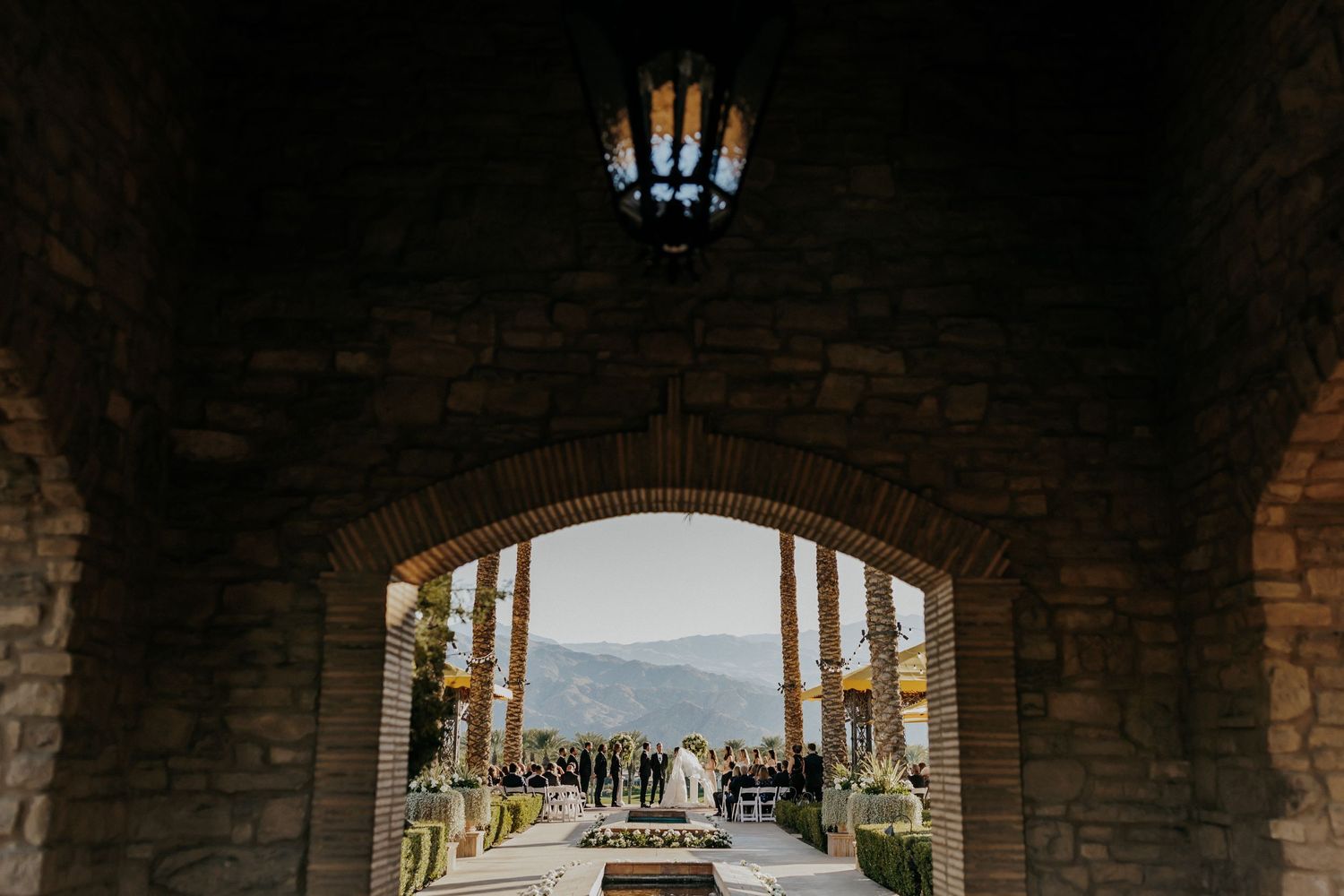 Stone archway entrance at a mountain venue frames guests gathering for a wedding ceremony in the distance.