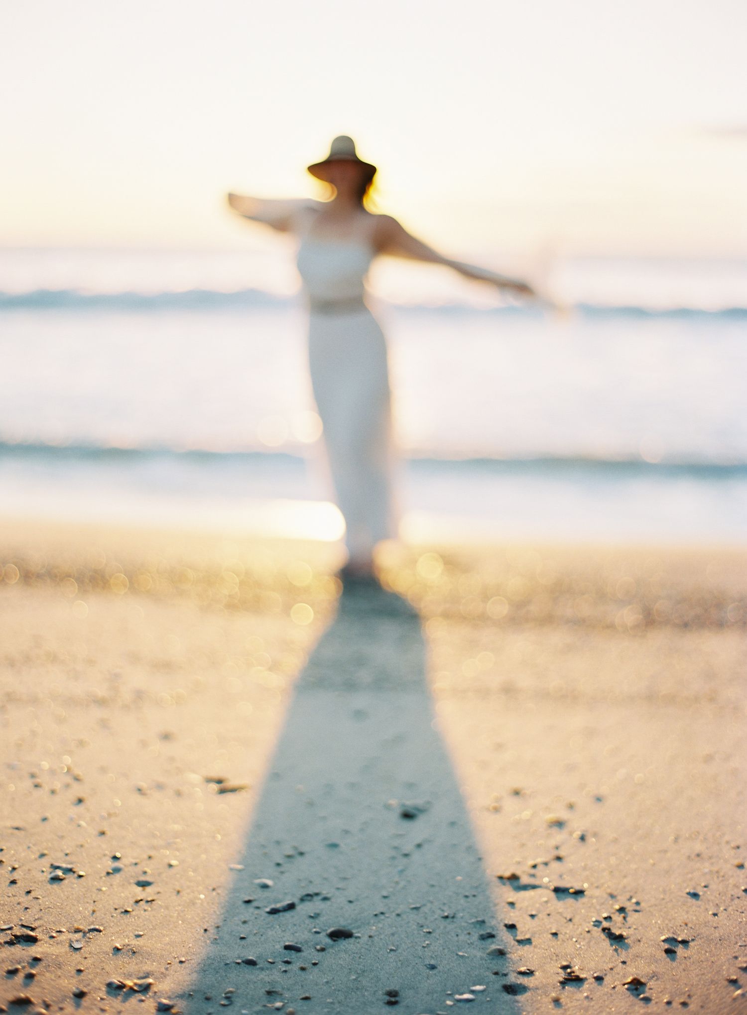 Figure in white dress and sunhat stands with arms outstretched on beach at sunset casting long shadow.
