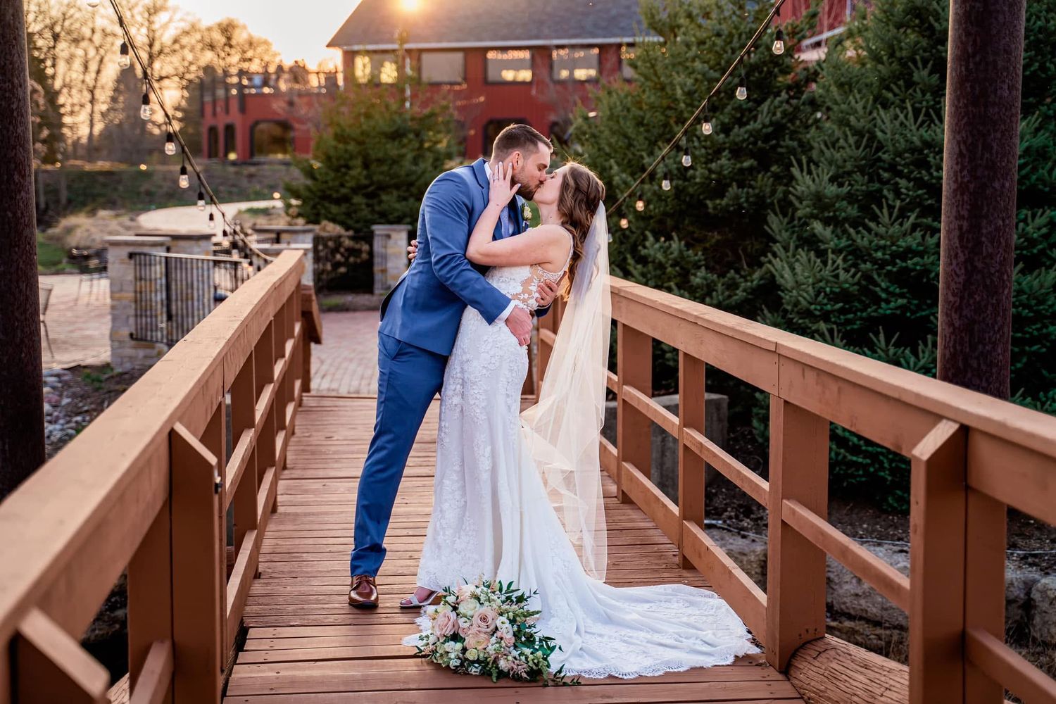 A couple shares a romantic kiss on a wooden bridge during sunset with a bouquet at their feet and red building in background.