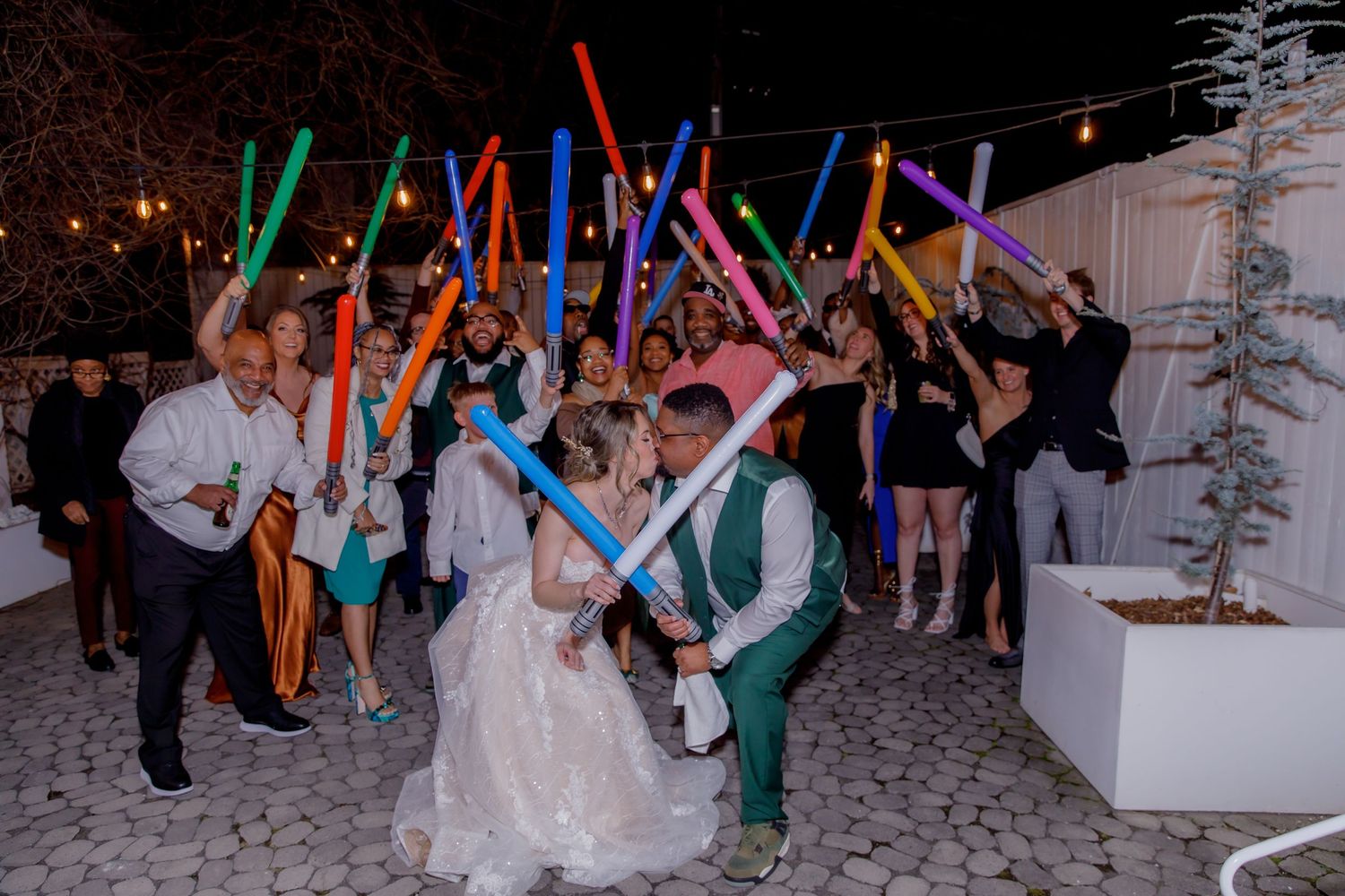 Wedding party guests hold colorful light sabers while celebrating at an outdoor evening reception.