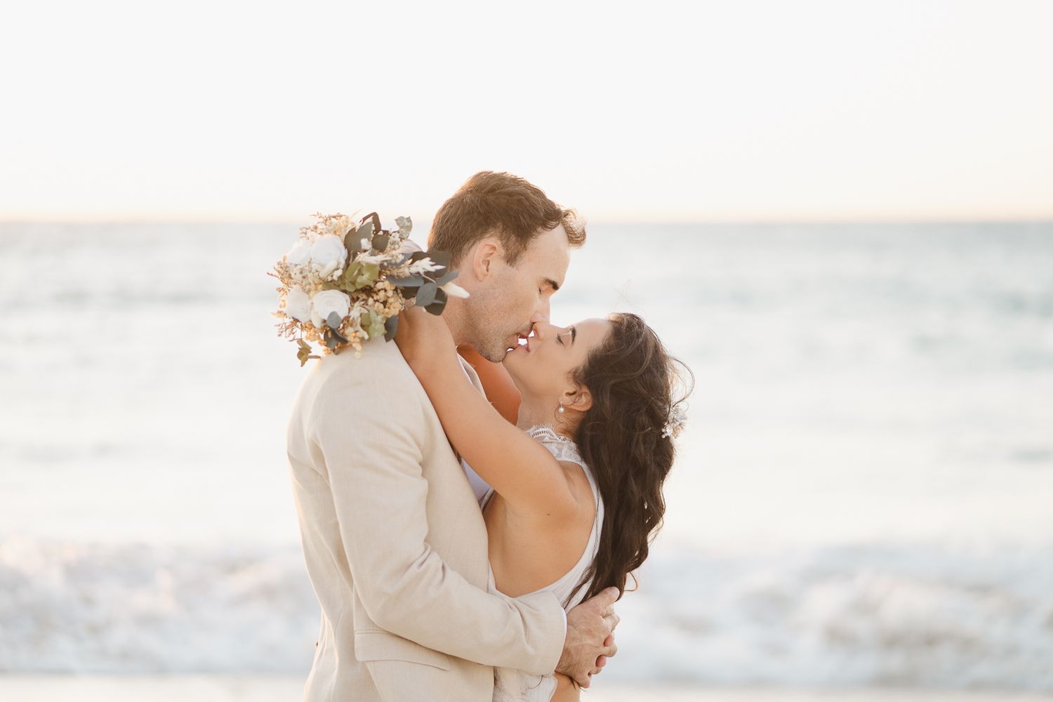 Romantic beach wedding scene with couple embracing by the ocean at sunset while bride holds floral bouquet.