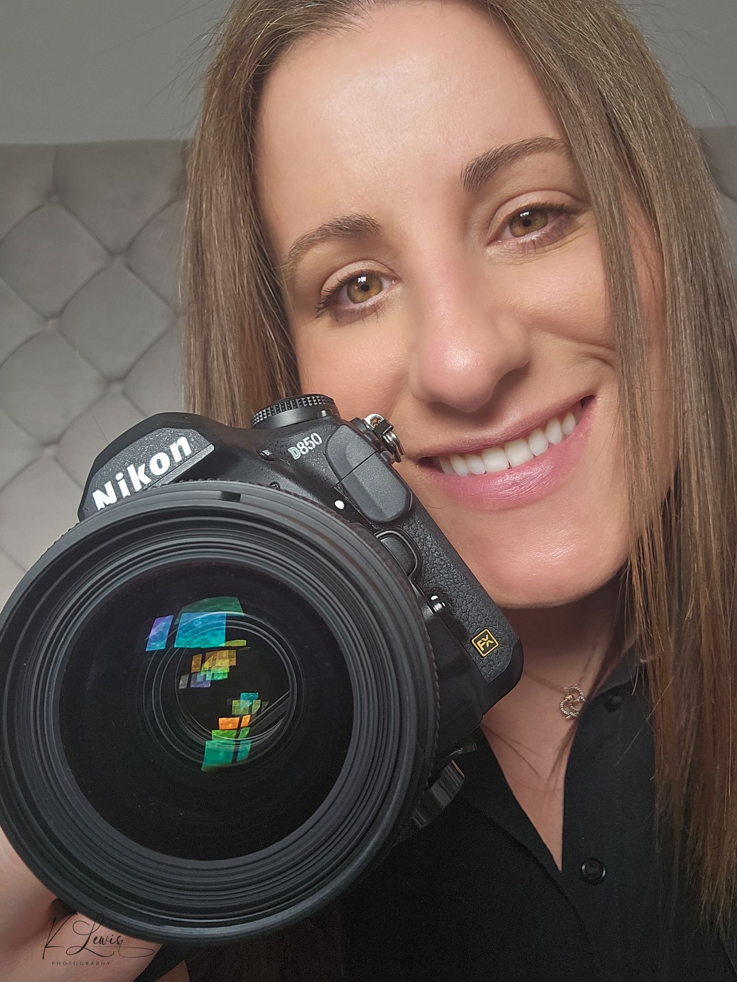 Smiling female photographer holding a Nikon D850 DSLR camera with colorful lens reflection in selfie portrait.
