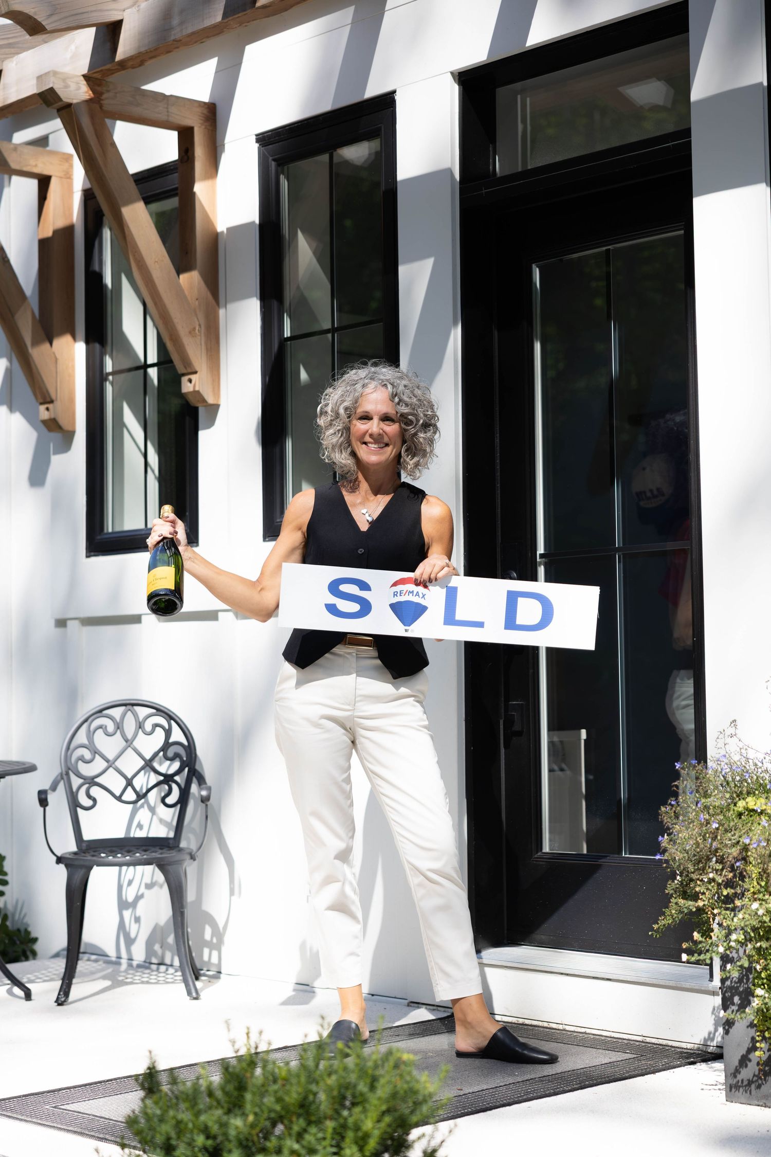 Smiling woman holding a SOLD sign and champagne bottle in front of a modern white home entrance.