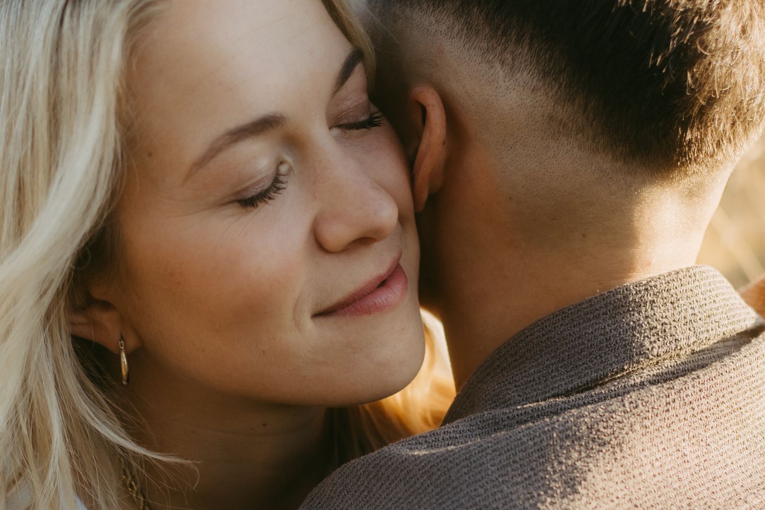 Blonde woman with eyes closed smiling softly as she nuzzles against a man in a brown jacket in warm golden light.