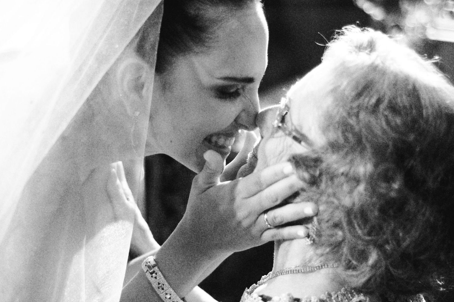 Black and white photo of a smiling bride in veil sharing a tender moment with an older woman at a wedding.